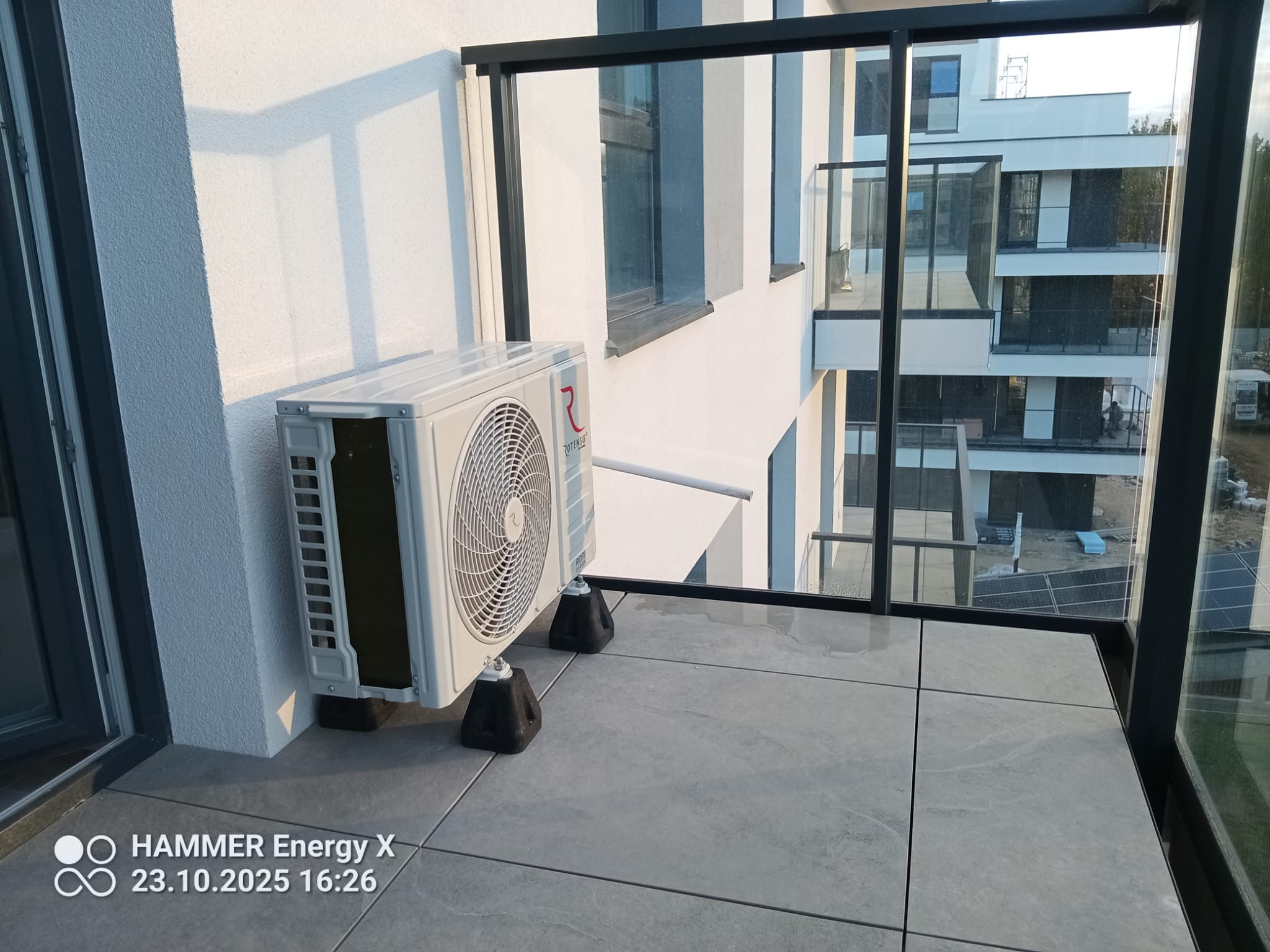 Biała jednostka zewnętrzna klimatyzacji Rotenso na balkonie nowoczesnego apartamentu, widok przez szklaną balustradę na sąsiedni budynek. Montaż na gumowych podkładkach antywibracyjnych.