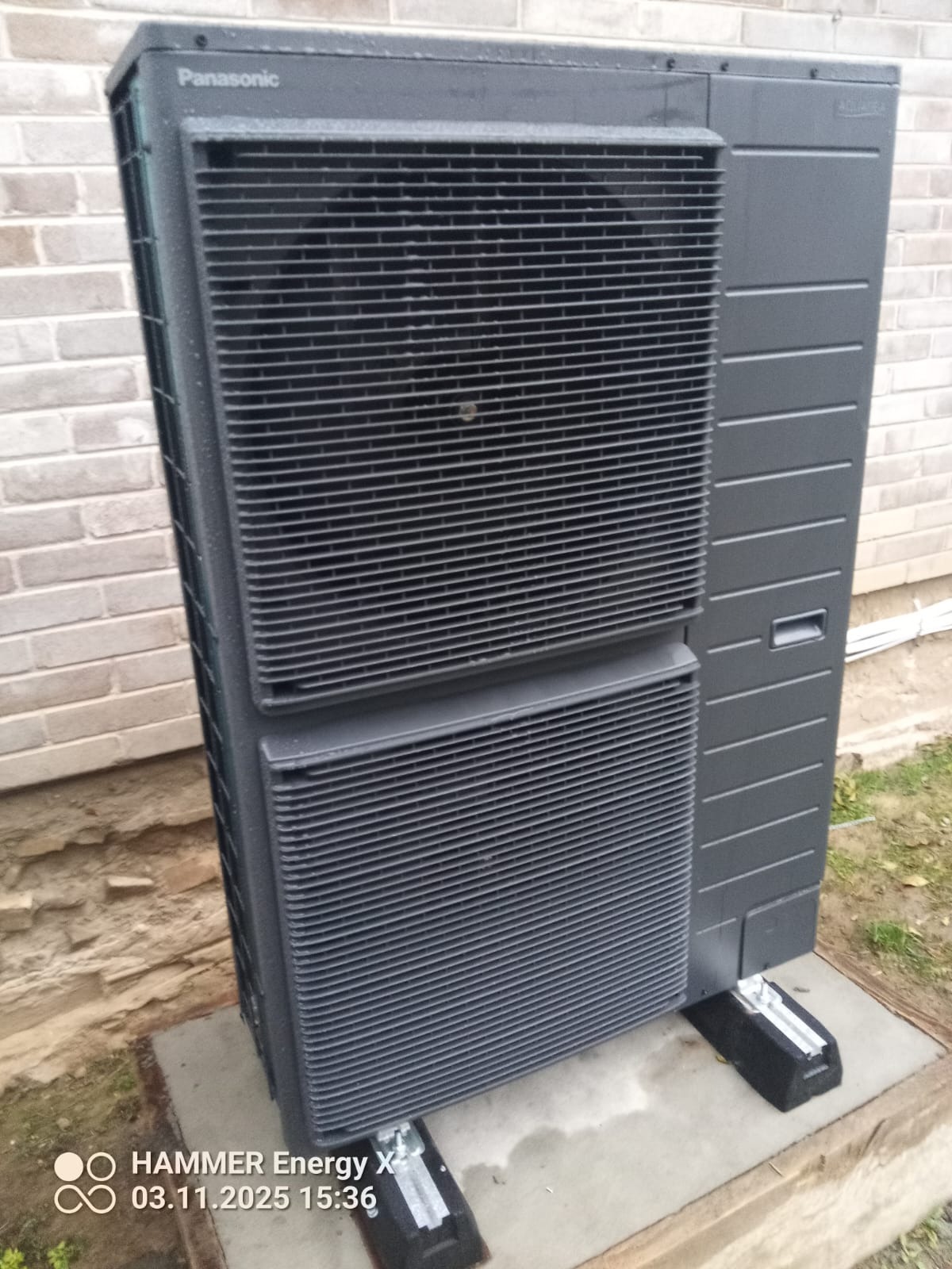 Zewnętrzna jednostka pompy ciepła Panasonic Aquarea, zamontowana na betonowej podstawie przy ceglanym murze. Widoczne detale konstrukcyjne i logo producenta.