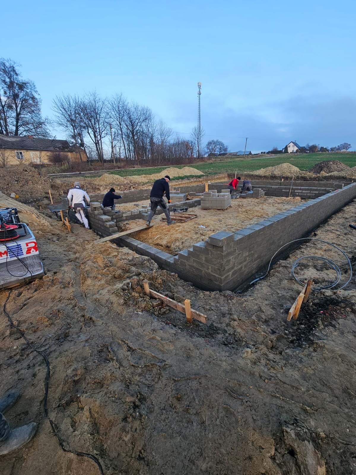 Budowa fundamentów domu z bloczków betonowych. Widoczni pracownicy w trakcie murowania ścian fundamentowych. Na pierwszym planie wykop i ziemia.