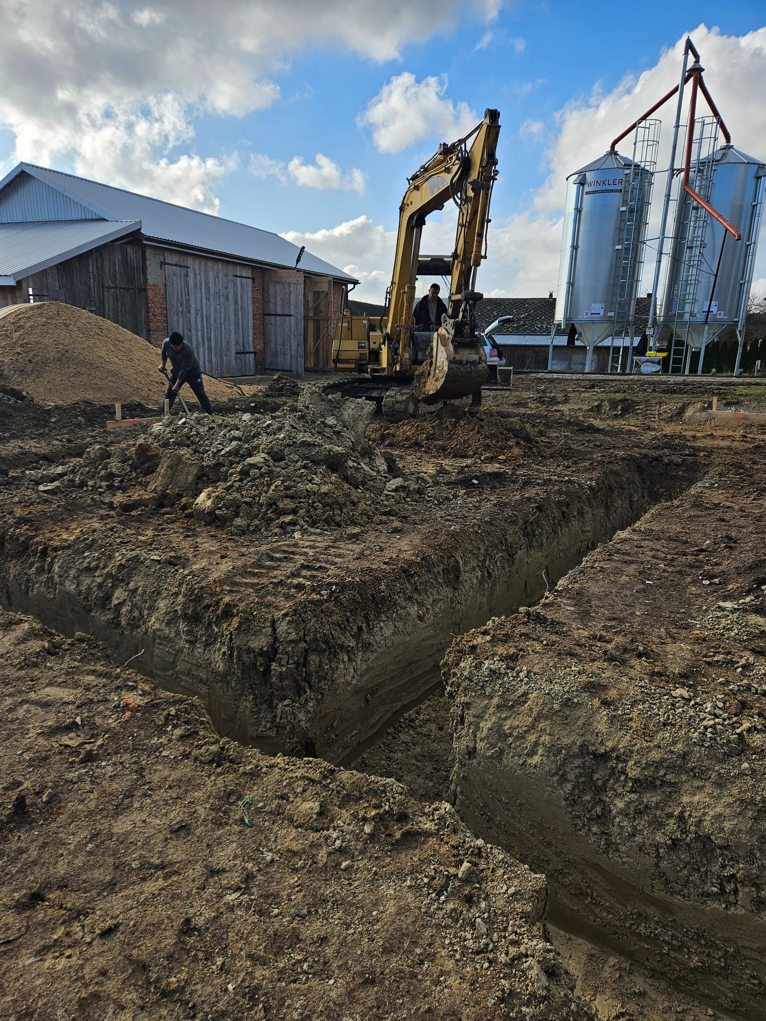 Wykop pod fundamenty budynku gospodarczego z użyciem koparki i pracy ręcznej, widoczne silosy zbożowe i budynek w tle, błękitne niebo z chmurami.