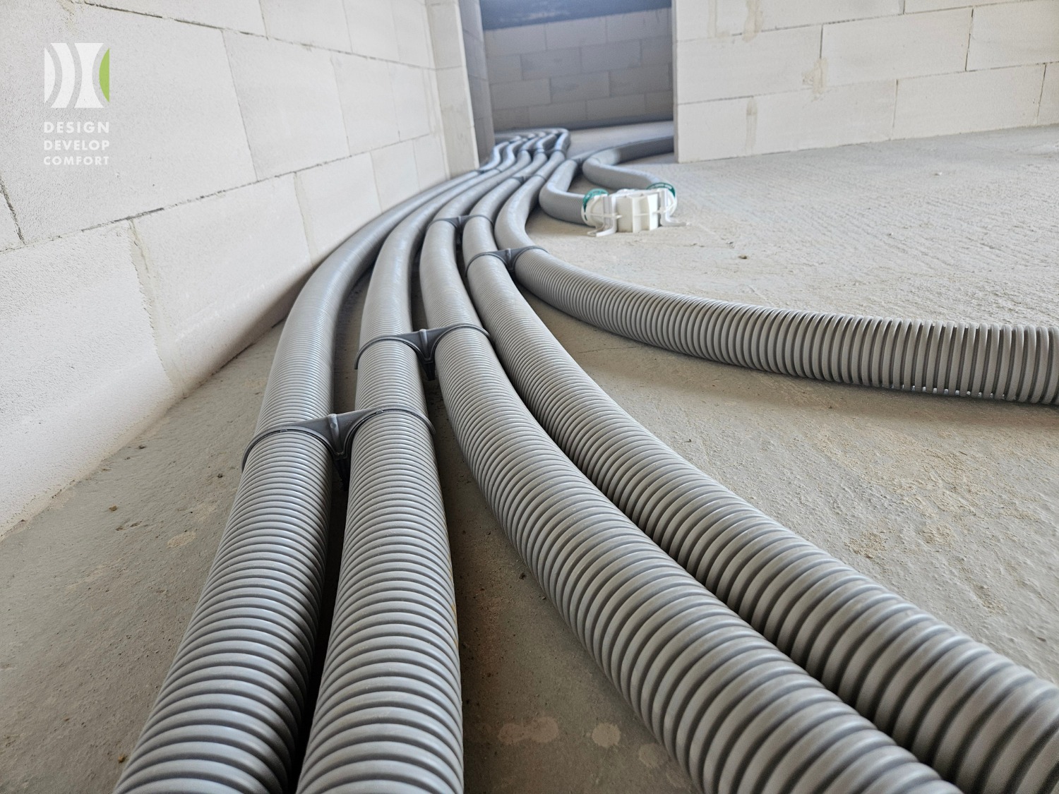 Szereg szarych, karbowanych rur wentylacyjnych ułożonych równolegle na surowym betonie, przymocowanych do ściany z białych bloczków. Widok z niskiej perspektywy.