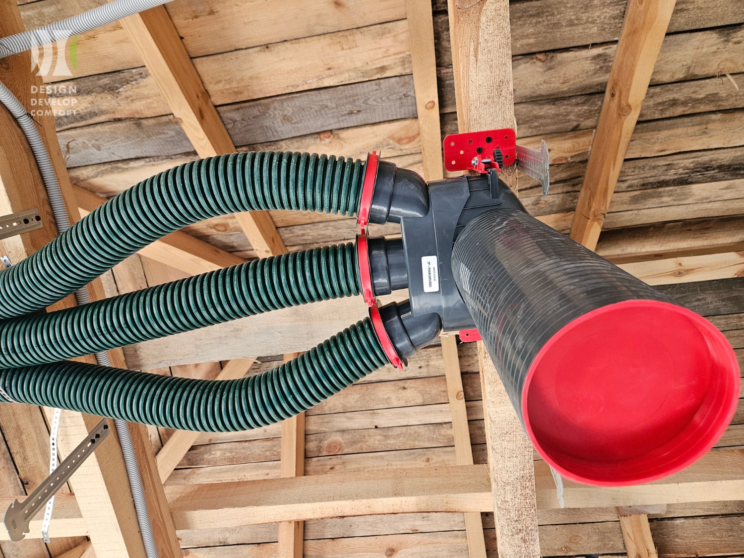 Rozgałęziona instalacja wentylacyjna z zielonymi, elastycznymi przewodami w surowym wnętrzu poddasza, widok z dołu, z elementami montażowymi w kolorze czerwonym.