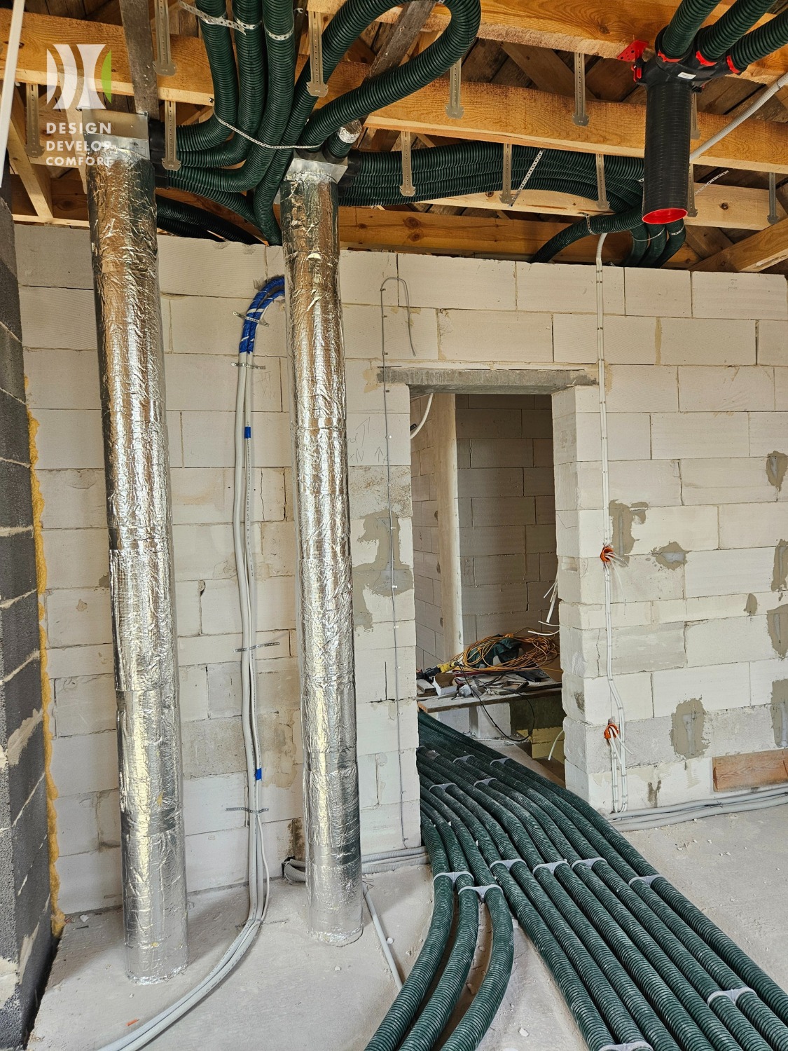 Montaż instalacji rekuperacji w surowym wnętrzu: zielone rury wentylacyjne, srebrne izolowane przewody, ściany z białego bloczka, widoczne elementy konstrukcji dachu.