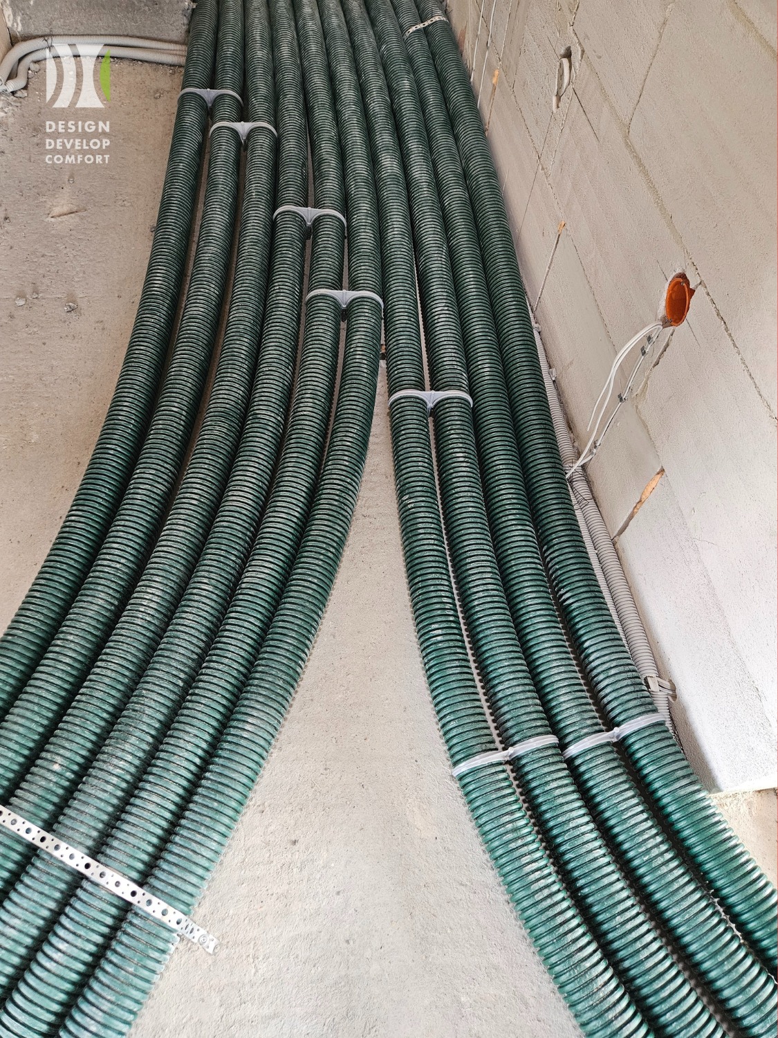 Zielone, karbowane rury wentylacyjne ułożone równolegle na podłodze w surowym wnętrzu, przymocowane uchwytami. Widoczna instalacja elektryczna w ścianie.