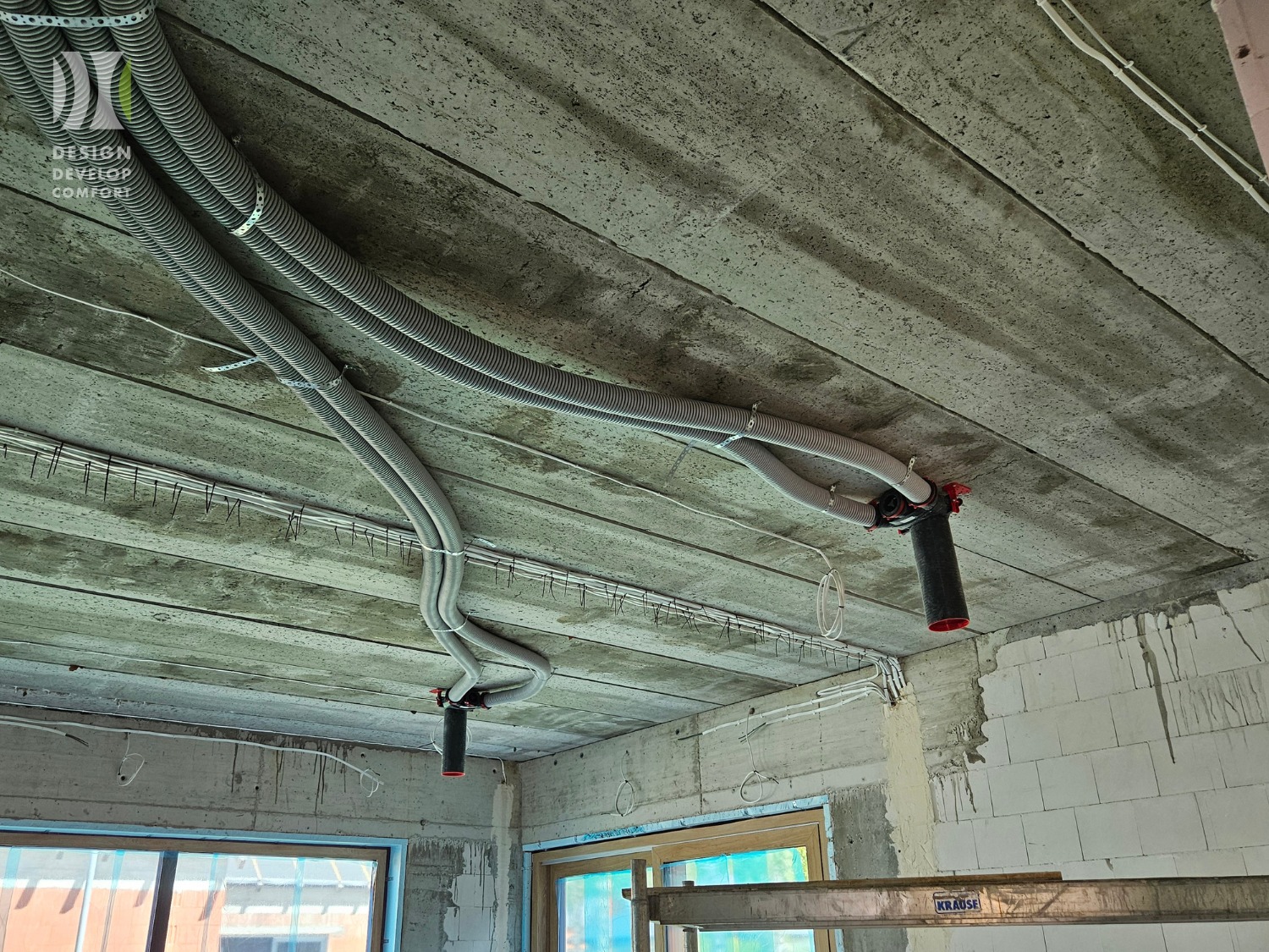 Szare, karbowane rury wentylacyjne pod betonowym stropem, zamocowane do czarnych, cylindrycznych elementów. Widoczne okablowanie i fragment ściany z pustaków.