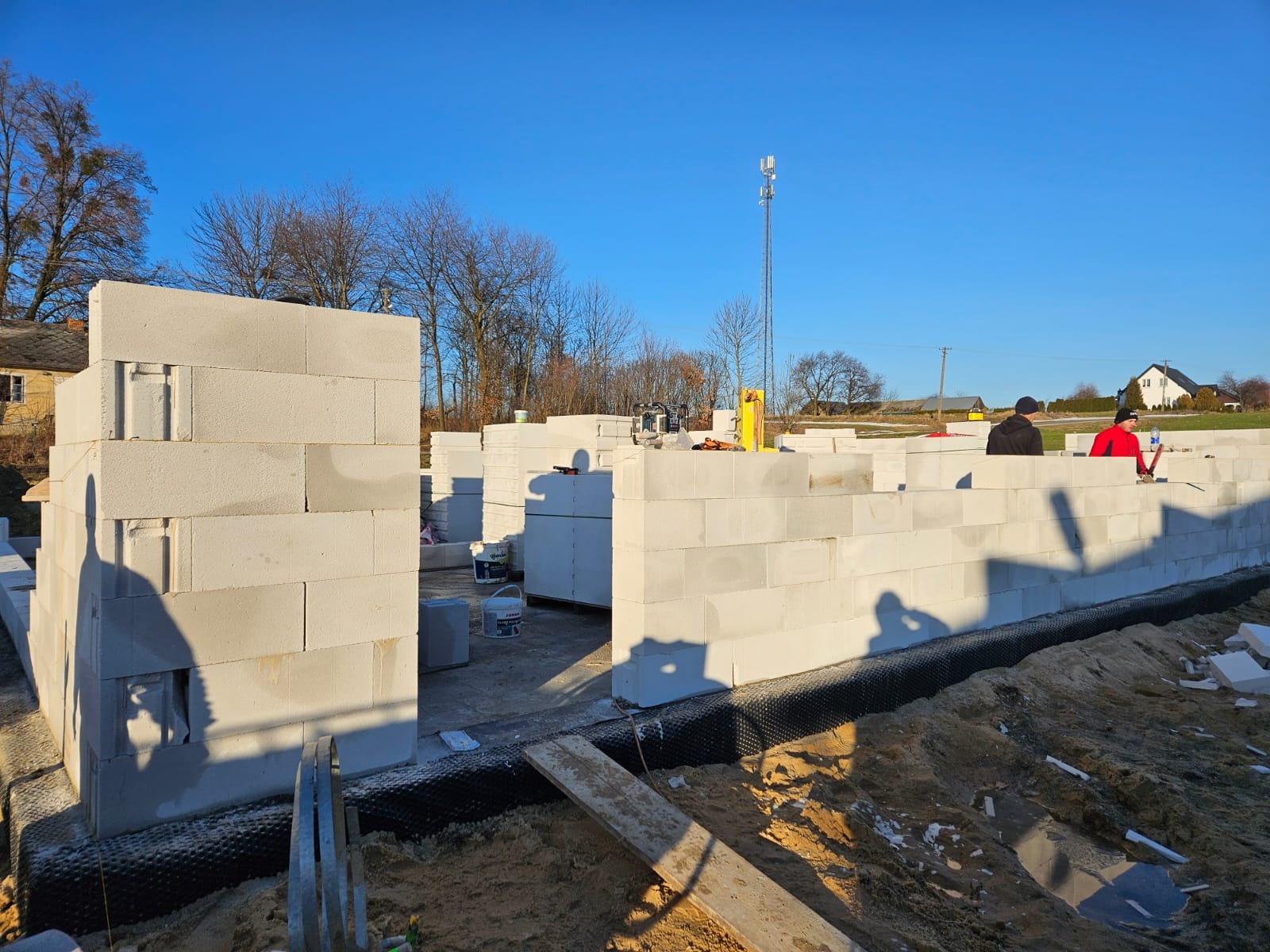 Budowa domu z betonu komórkowego: wzniesione ściany fundamentowe z izolacją, widoczne narzędzia i ekipa budowlana w słoneczny dzień. Etap początkowy budowy.