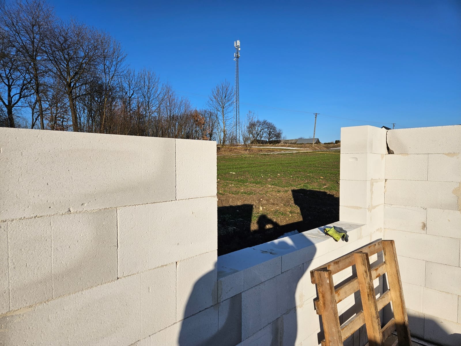 Wznoszenie ściany z białych bloczków betonu komórkowego Ytong. Widok przez otwór okienny na pole i wieżę telekomunikacyjną. Paleta i rękawice robocze obok.