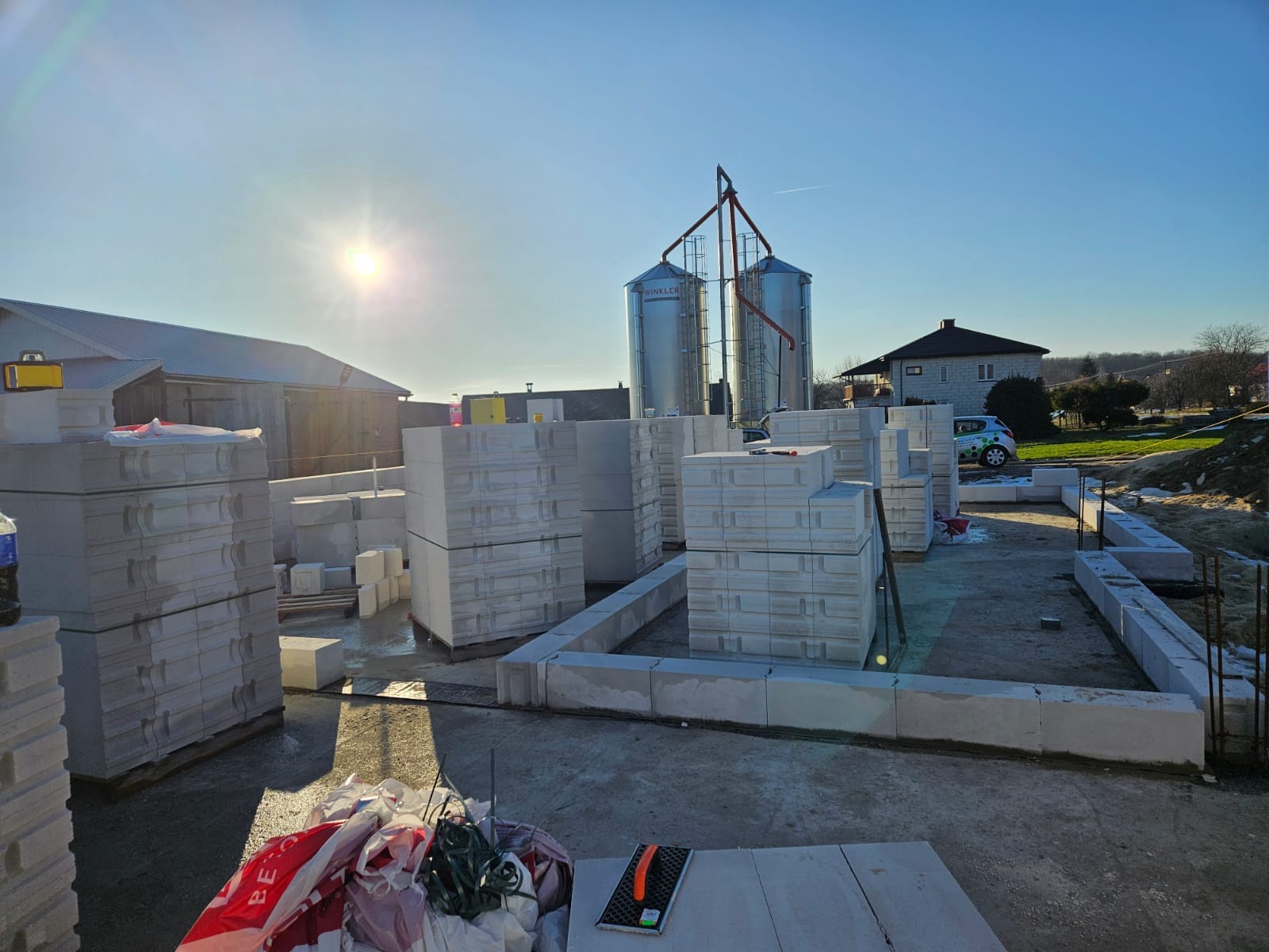 Budowa fundamentów z bloczków betonu komórkowego, widoczne silosy w tle, słoneczny dzień. Etap wznoszenia ścian fundamentowych.