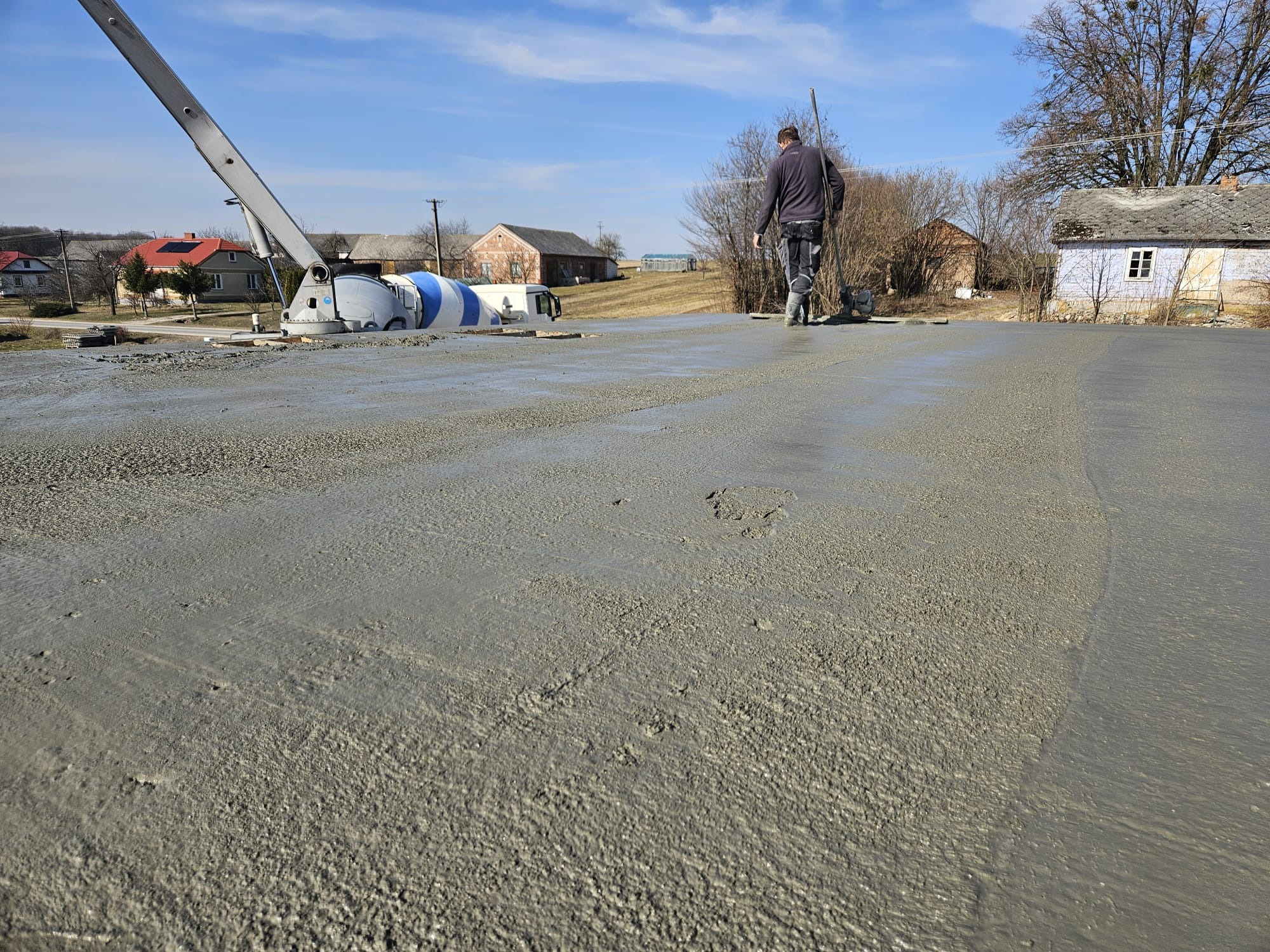 Wylewanie stropu z betonomieszarki na budowie domu jednorodzinnego. Pracownik wyrównuje powierzchnię. W tle zabudowa wiejska i błękitne niebo.