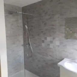 Wykończona łazienka z kabiną prysznicową ze szklaną ścianką, szare płytki imitujące kamień z dekoracyjnym panelem mozaiki, biała umywalka z szafką, widok z niskiego kąta.