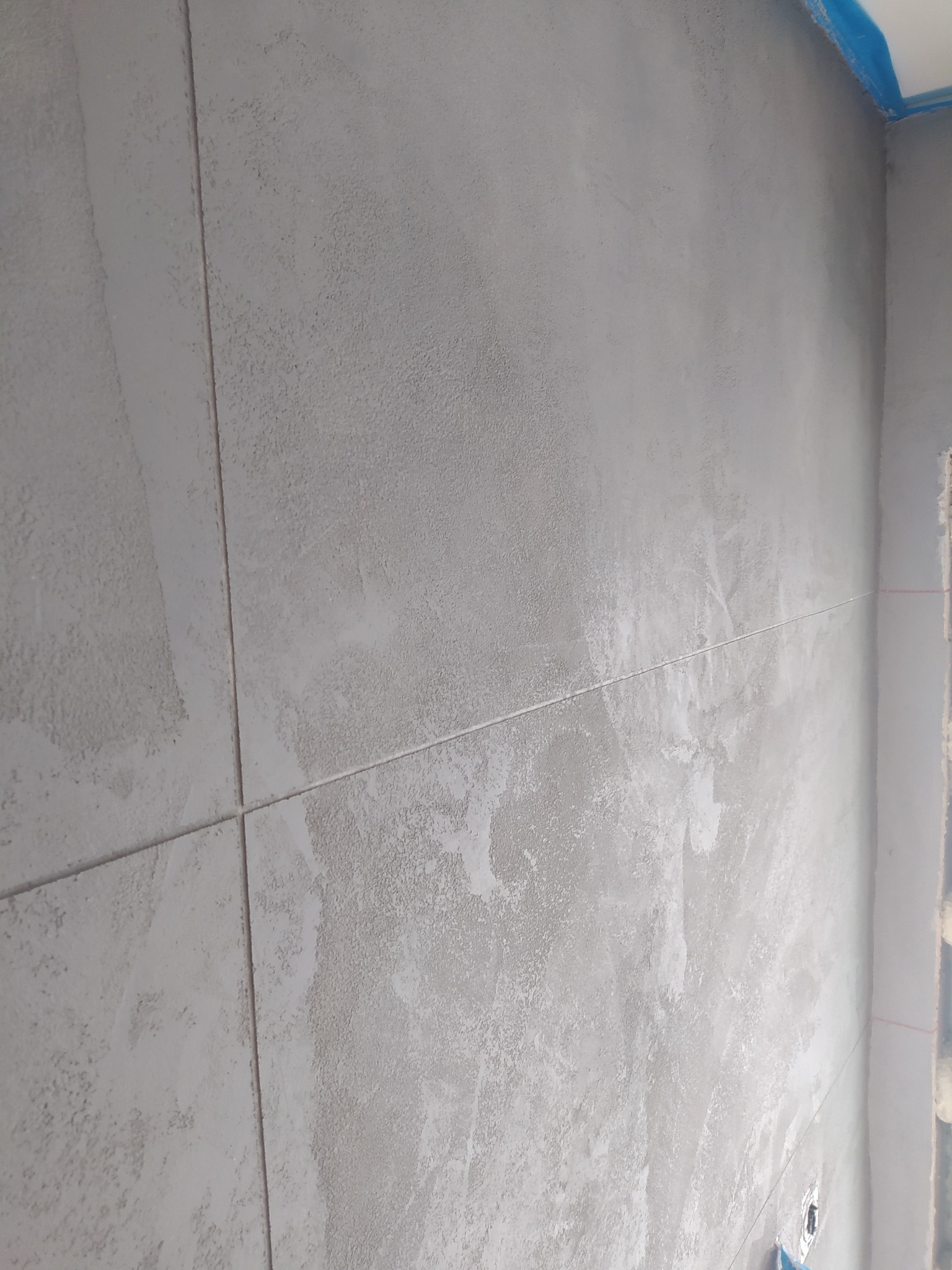 Szara ściana w łazience po nałożeniu tynku dekoracyjnego, widoczne łączenia płyt i niebieska taśma malarska przy suficie.