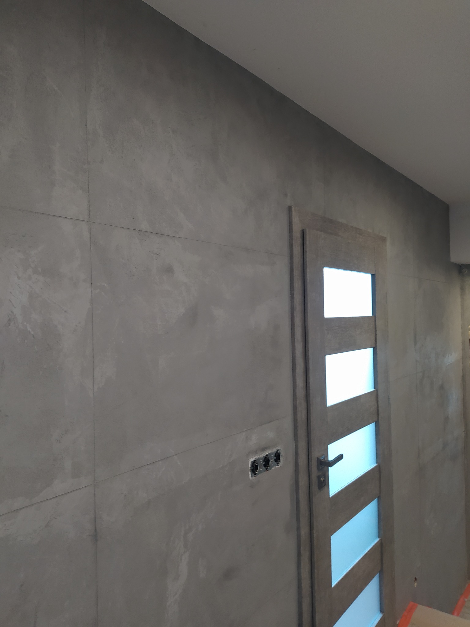Szara ściana wykończona płytami imitującymi beton, z widoczną drewnianą ościeżnicą i drzwiami z czterema przeszkleniami, gniazdka elektryczne bez obudowy.