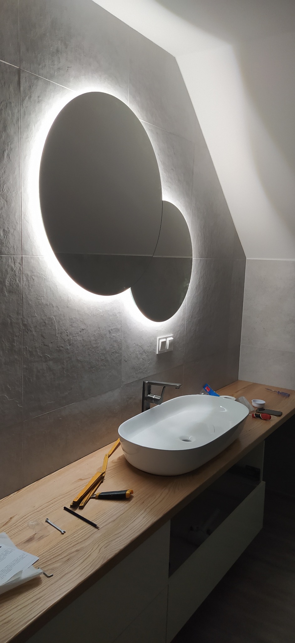 Nowoczesna łazienka w trakcie montażu: owalna umywalka nablatowa, bateria, podświetlane okrągłe lustro na szarych płytkach, drewniany blat, narzędzia na blacie, skośny sufit.