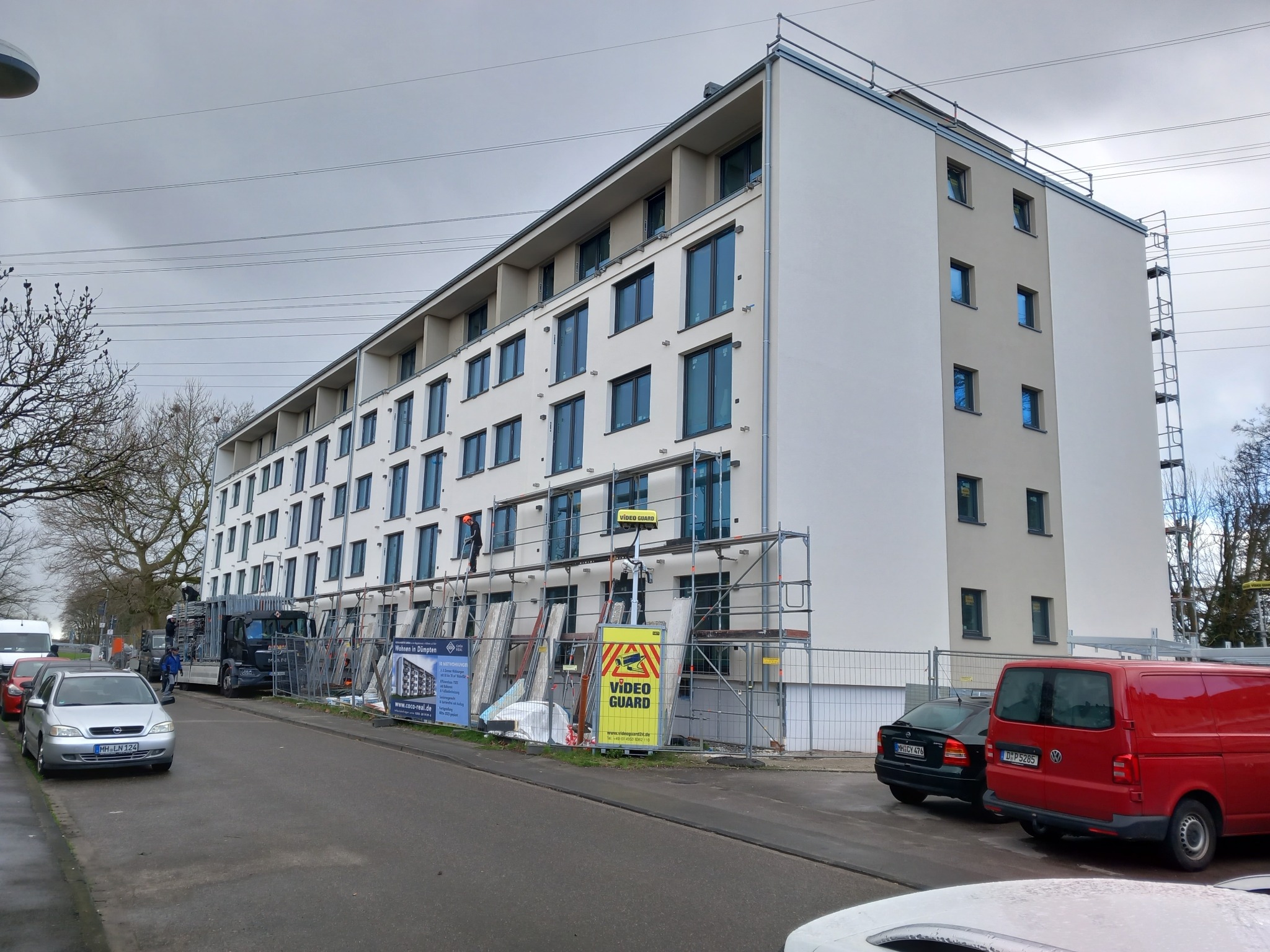 Wielopiętrowy budynek w trakcie prac elewacyjnych, widoczne rusztowania, materiały budowlane i pracownicy na rusztowaniach, zaparkowane samochody na ulicy.