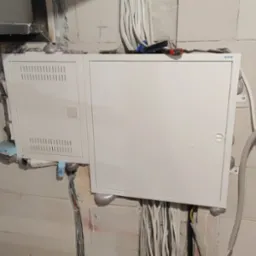 Dwa białe panele elektryczne zamontowane na ścianie z bloczków betonowych, uszczelnione pianką montażową, z widocznymi kablami i narzędziami na górze.