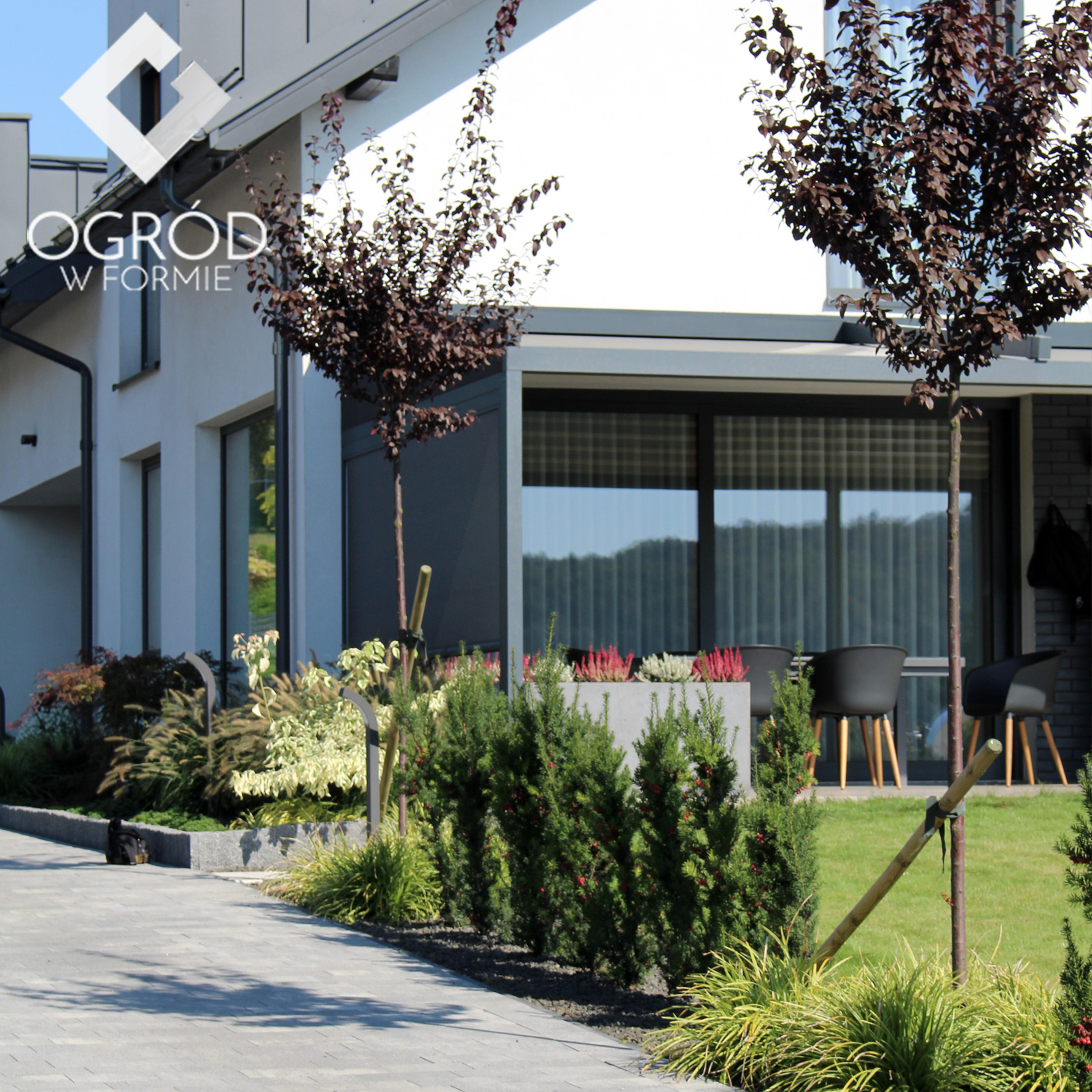 Nowoczesny ogród przydomowy z ścieżką z kostki, niskimi krzewami iglastymi z czerwonymi owocami, purpurowymi drzewkami, minimalistycznym tarasem z szarymi meblami i logotypem firmy ogrodniczej...