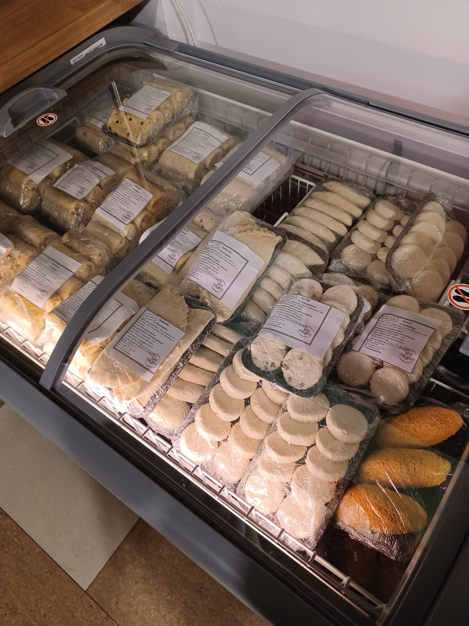 Chłodnia z różnymi rodzajami mrożonych dań gotowych: czebureki z twarogiem, serniki z rodzynkami i serniki z czekoladą, wszystkie zapakowane w folię z etykietami.