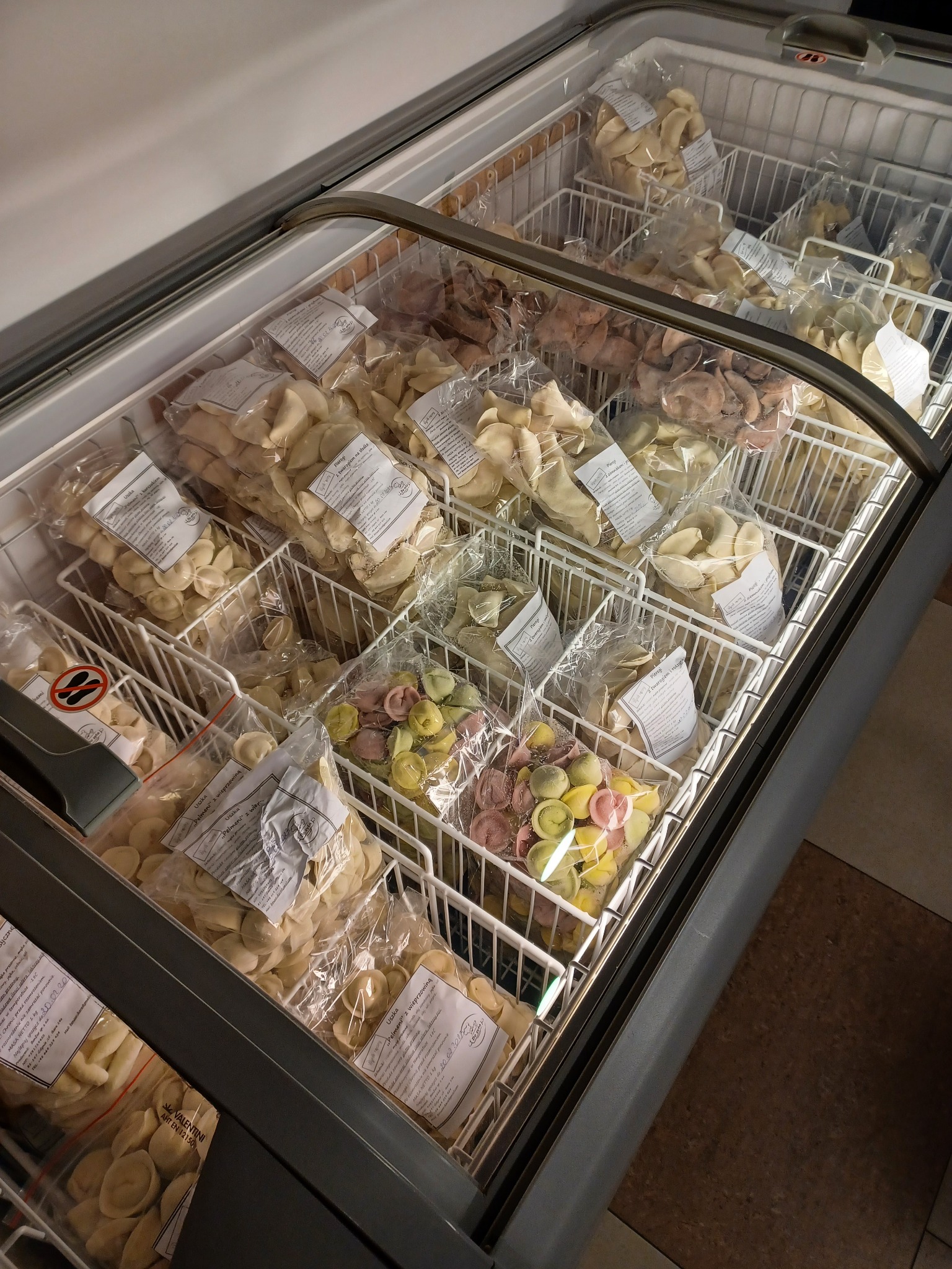 Zamrażarka sklepowa wypełniona różnymi rodzajami mrożonych pierogów i uszek w plastikowych opakowaniach, ułożonych w metalowych koszykach, niektóre z kolorowymi farszami.
