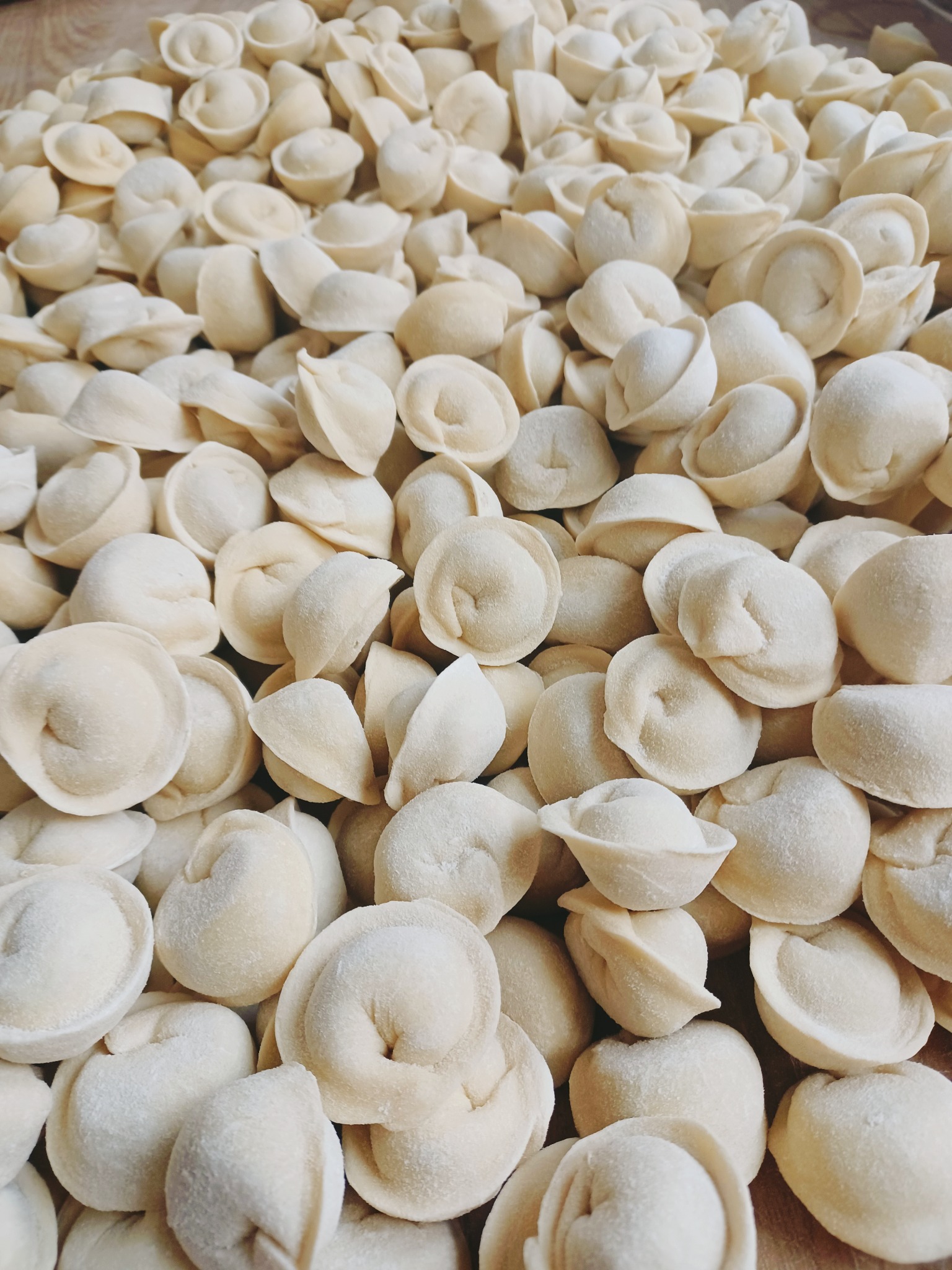 Góra surowych, ręcznie robionych pierożków (pelmeni) z delikatnym oprószeniem mąką, ułożonych na drewnianej powierzchni, gotowych do ugotowania.