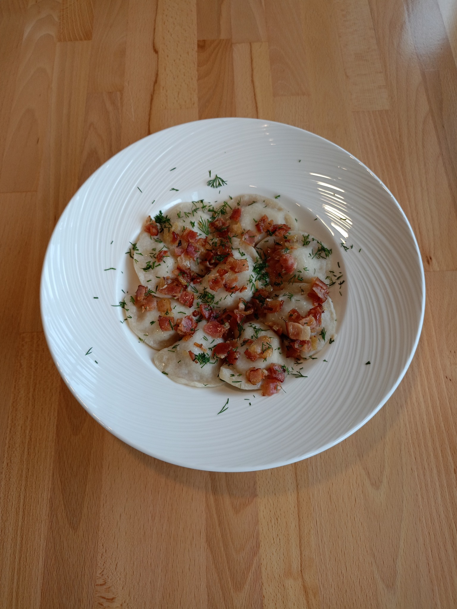 Porcja pierogów okraszonych skwarkami i koperkiem na białym, karbowanym talerzu, ustawiona na drewnianym blacie