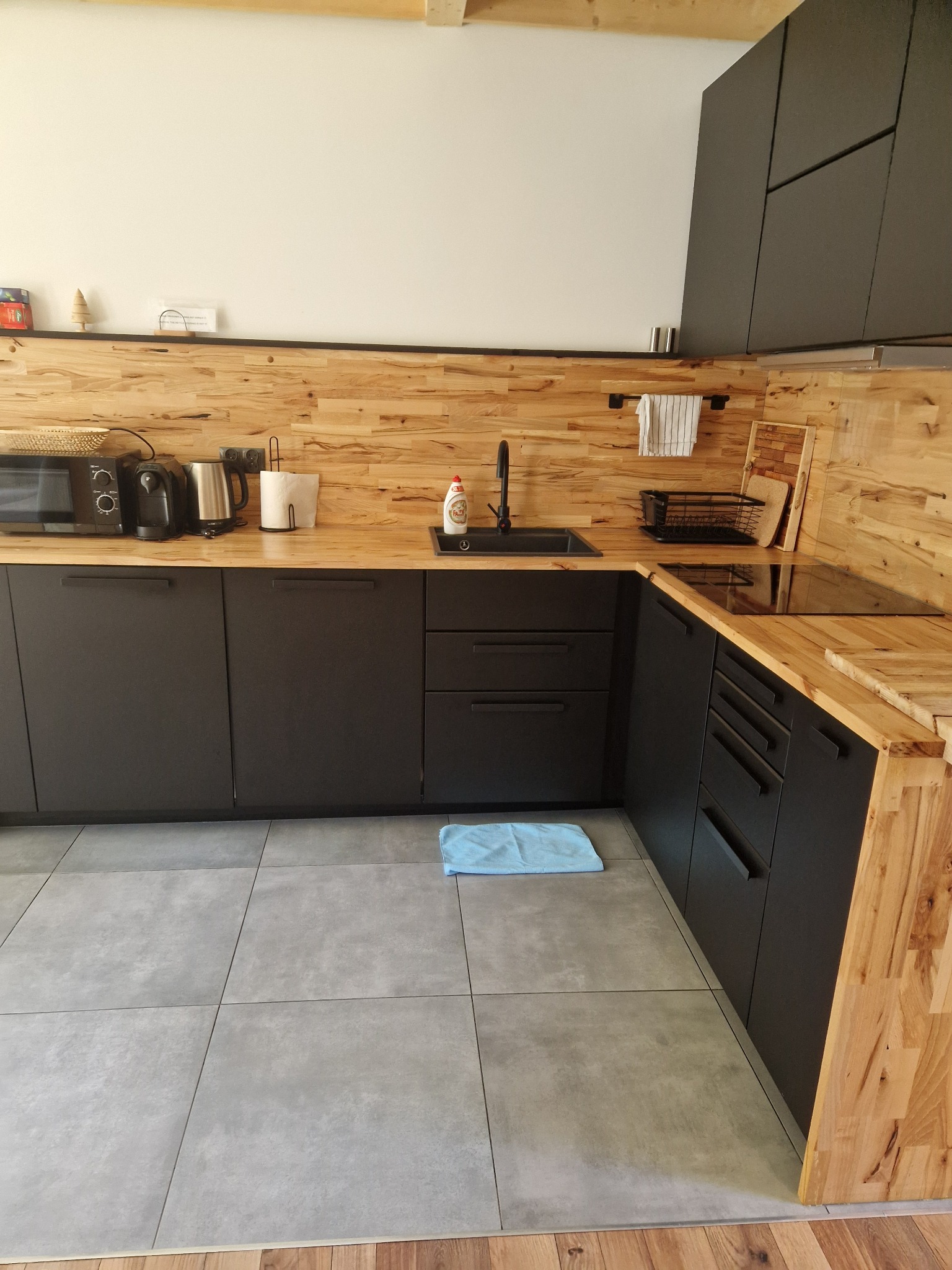 Nowoczesna kuchnia z czarnymi szafkami i drewnianym blatem, widoczny zlewozmywak, płyta indukcyjna, akcesoria kuchenne i niebieska ściereczka na podłodze.