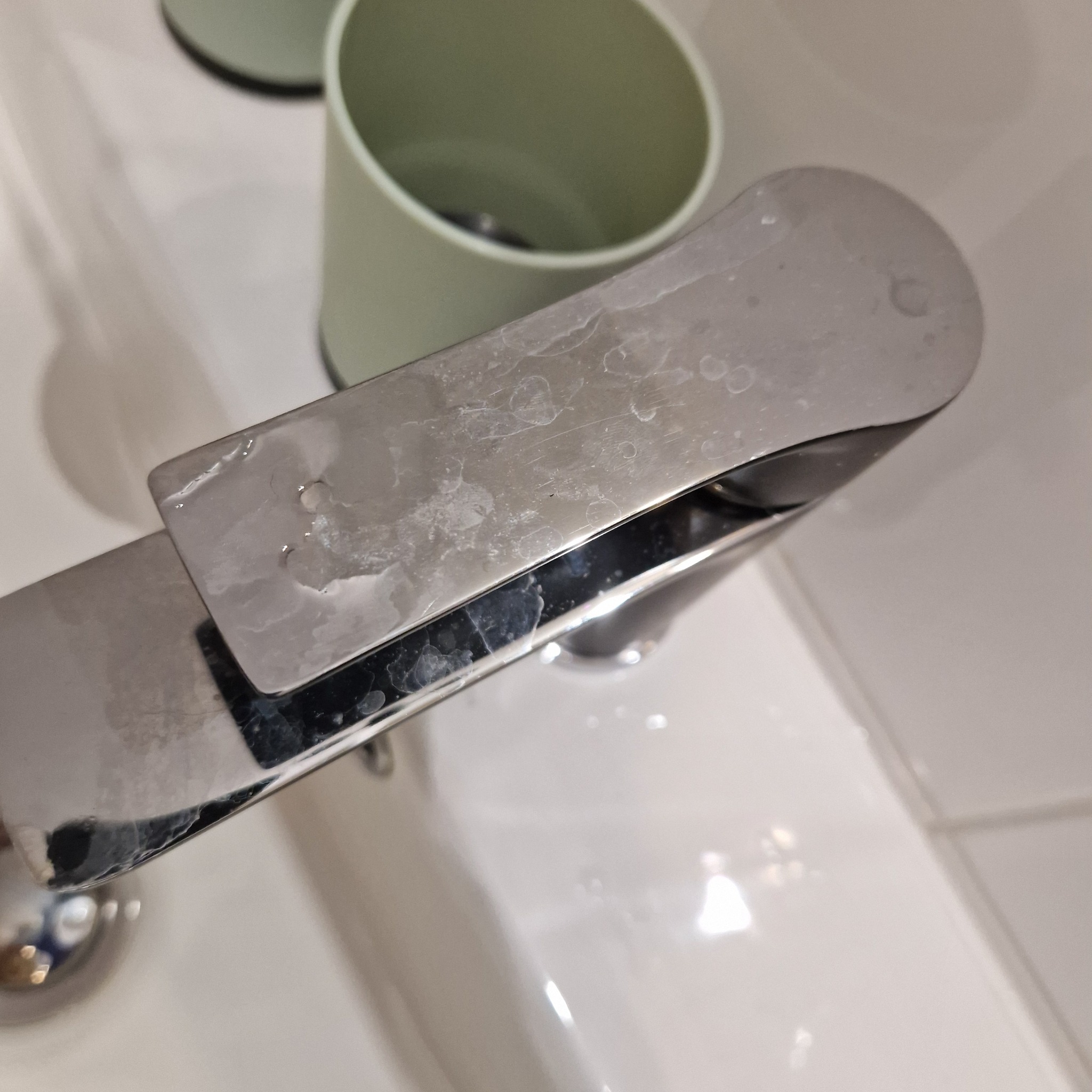 Bateria umywalkowa ze stali nierdzewnej pokryta osadem z kamienia, widoczne zacieki i matowa powierzchnia, obok kubek na szczoteczki do zębów.