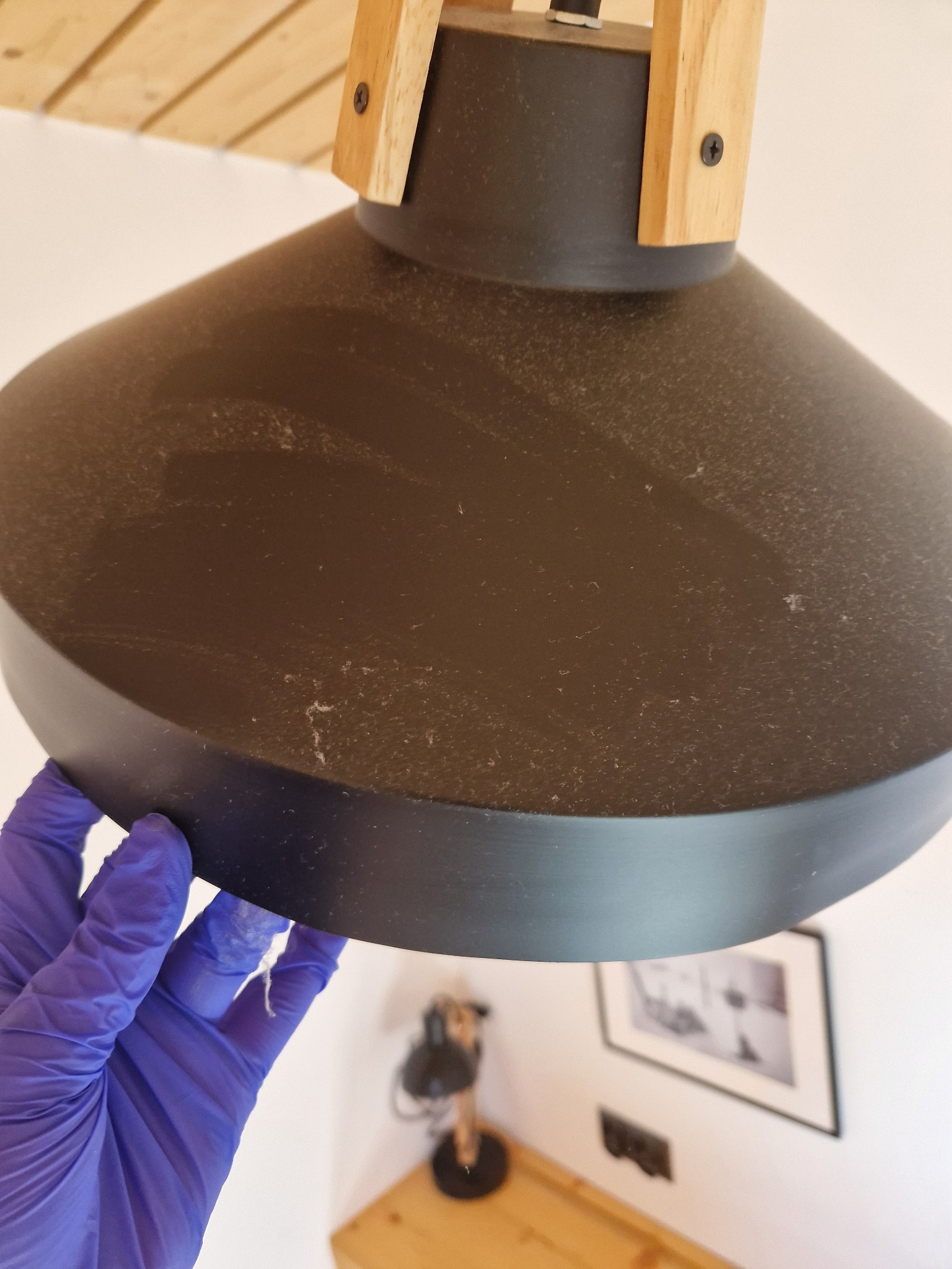 Ręka w niebieskiej rękawicy czyszcząca zakurzoną, czarną lampę z drewnianymi elementami w jasnym wnętrzu.