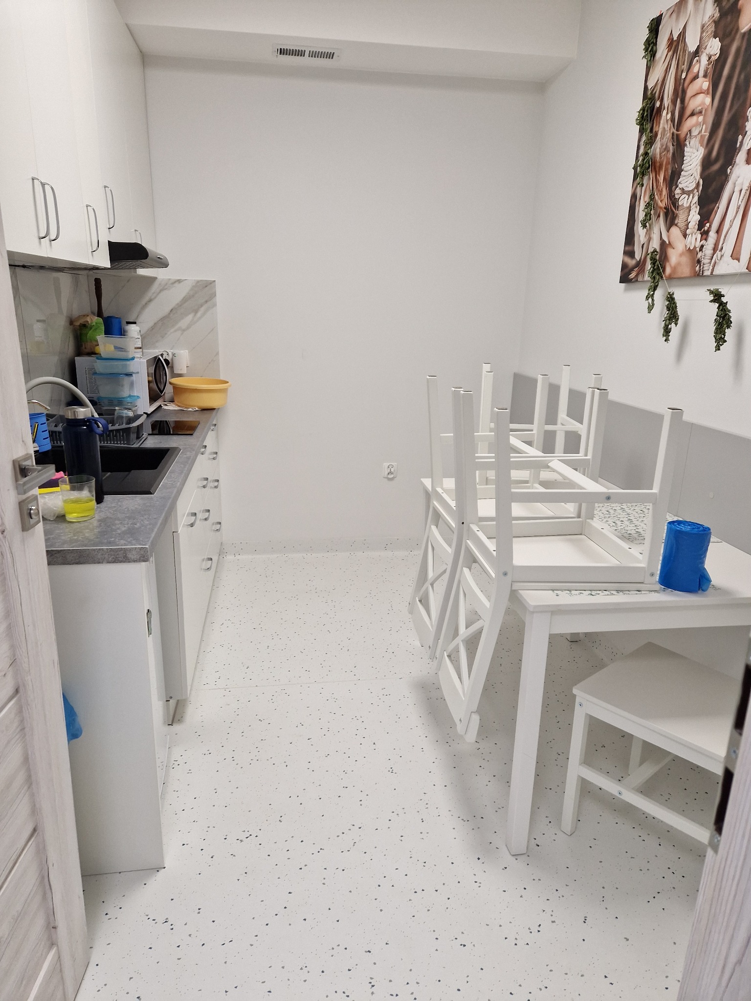 Jasne, minimalistyczne wnętrze z białymi meblami: szafki kuchenne, blat z umywalką, stół z krzesłami ułożonymi na blacie, na podłodze wzór w drobne kropki, na ścianie obraz.