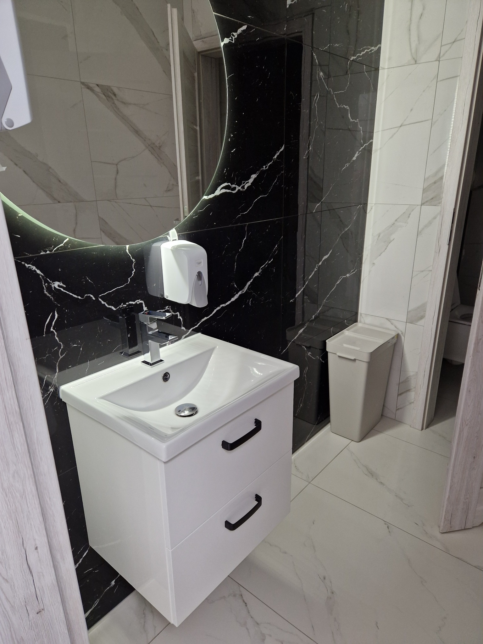 Nowoczesna biała umywalka z szafką i czarnymi uchwytami na tle marmurowej ściany z okrągłym lustrem z podświetleniem LED. Obok kosz na śmieci w kolorze beżowym.