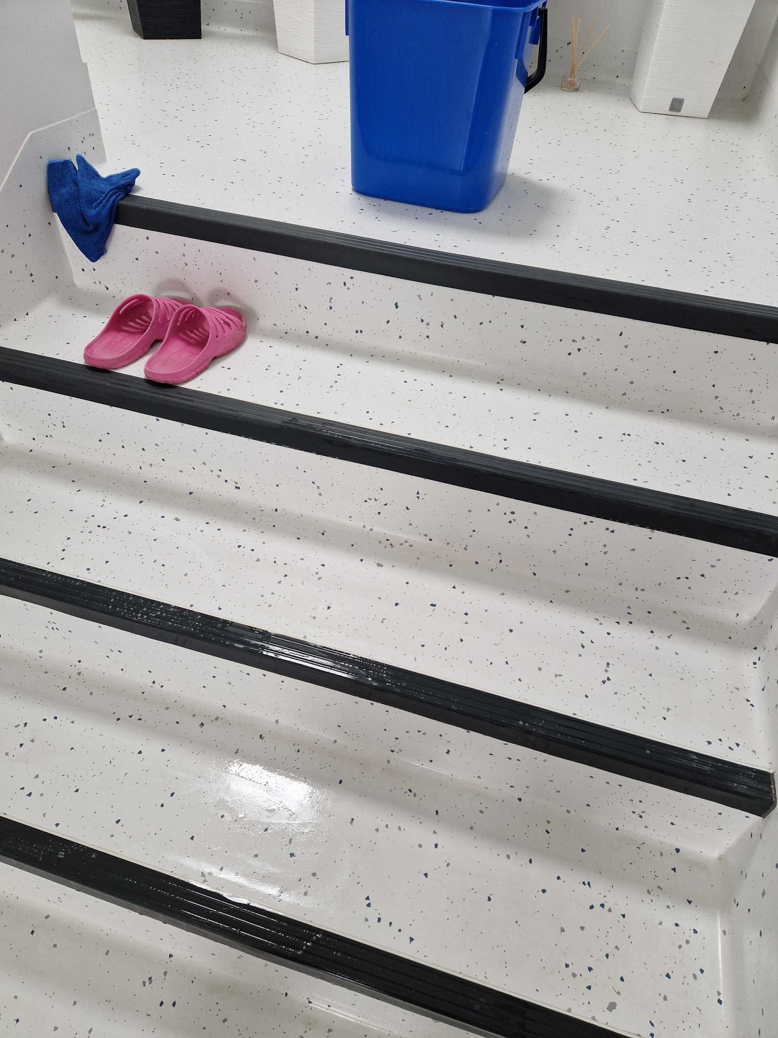 Białe schody z czarnymi paskami antypoślizgowymi, różowe klapki, niebieska ścierka i niebieskie wiadro na górnym stopniu. Podłoga z szarymi i niebieskimi plamkami.