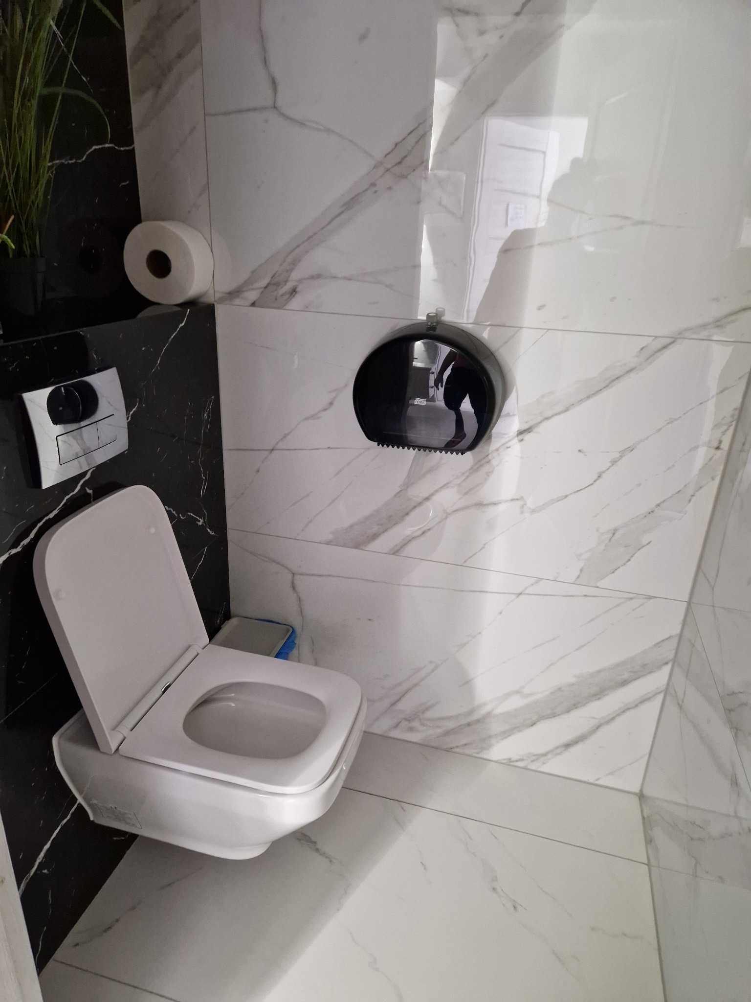 Nowoczesna toaleta z otwartą deską, czarno-białe marmurowe płytki, podajnik na ręczniki papierowe z odbiciem osoby.