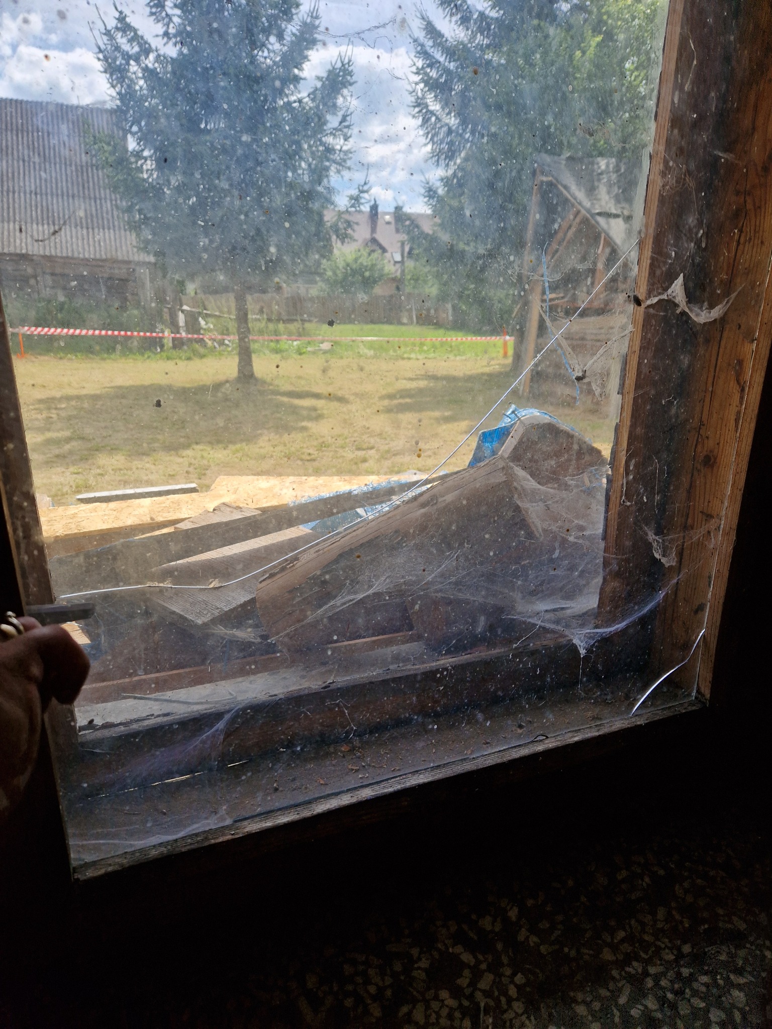 Brudna szyba okienna z pajęczynami, przez którą widać składowane deski, podwórko i drzewa w tle.