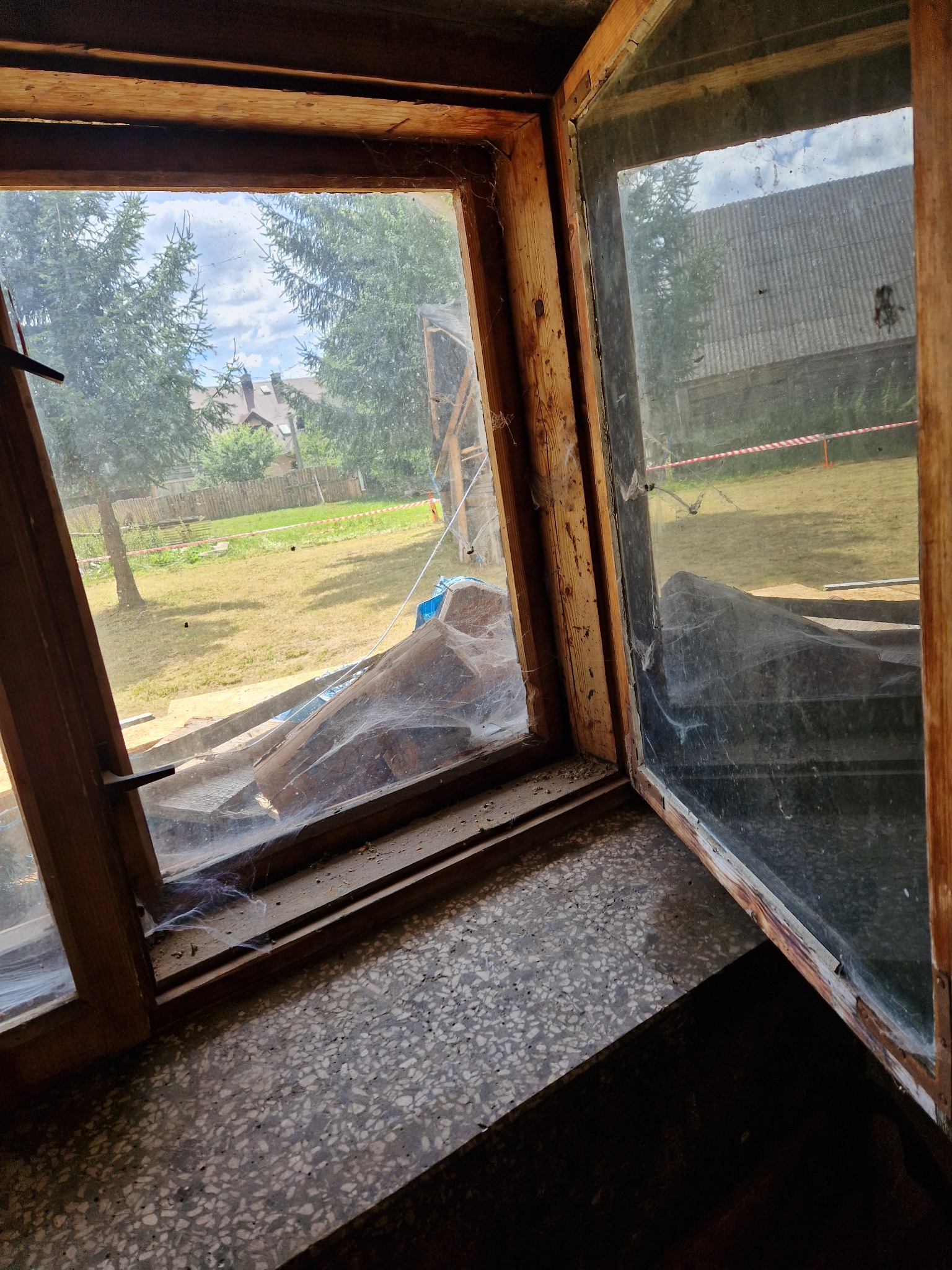 Otwarty drewniany okno z widocznymi pajęczynami na szybie i ramie, widok na zewnątrz na trawnik, drzewa i budynek z czerwono-białą taśmą ostrzegawczą.