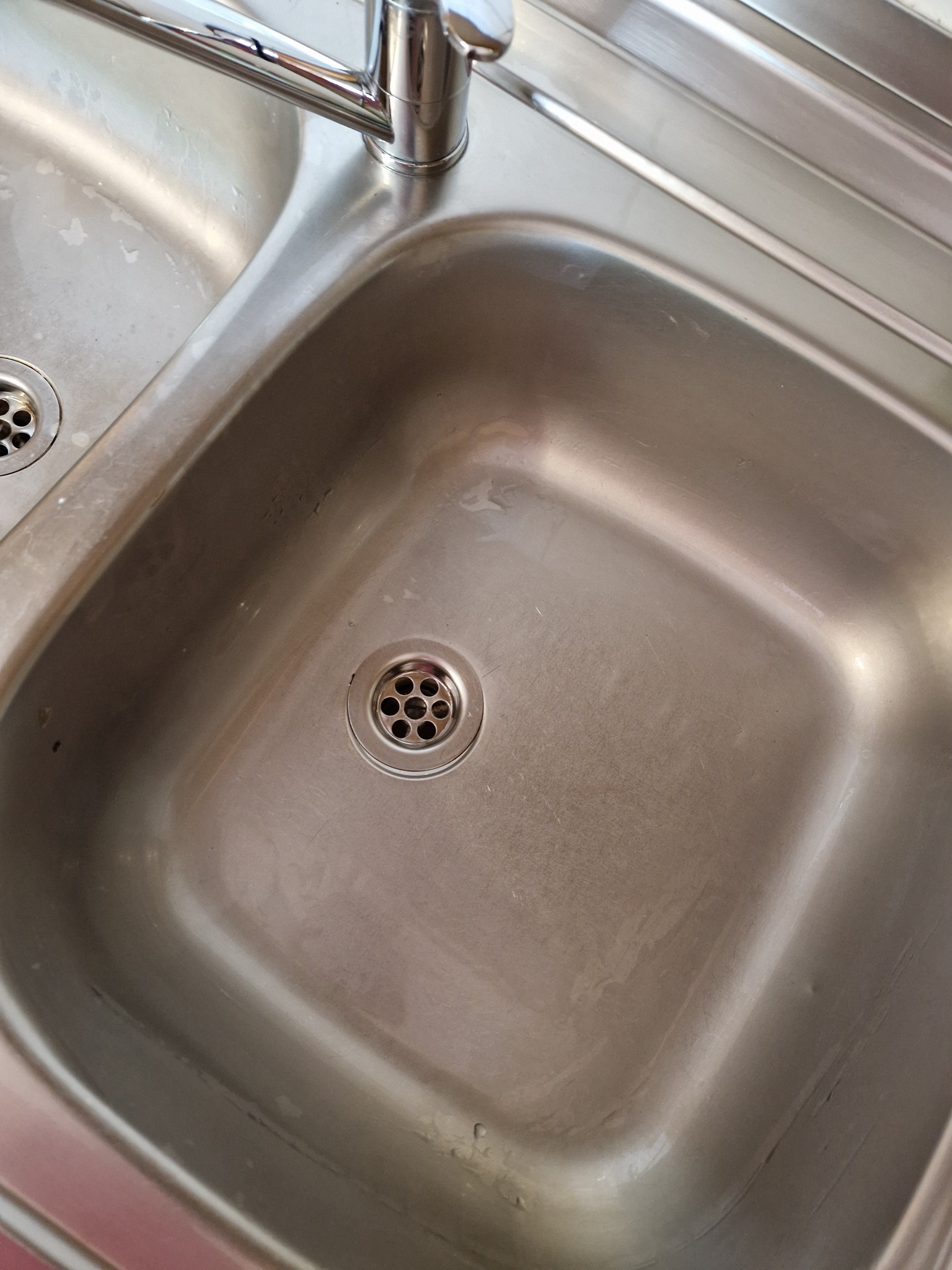 Metalowy, dwukomorowy zlew kuchenny ze stali nierdzewnej, widoczne ślady użytkowania i drobne zabrudzenia, kran w górnej części kadru.