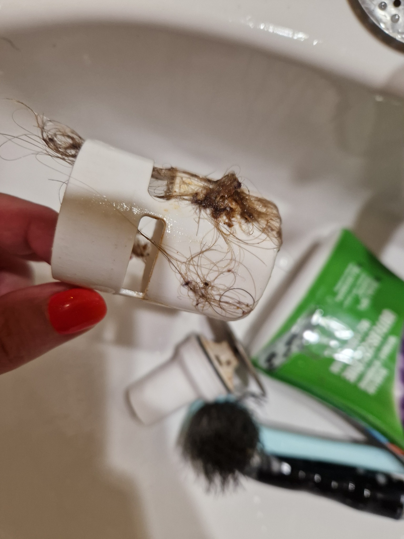 Zatkanie odpływu umywalki włosami w plastikowej obudowie, widoczna część dłoni z czerwonym paznokciem, w tle kosmetyki w umywalce.