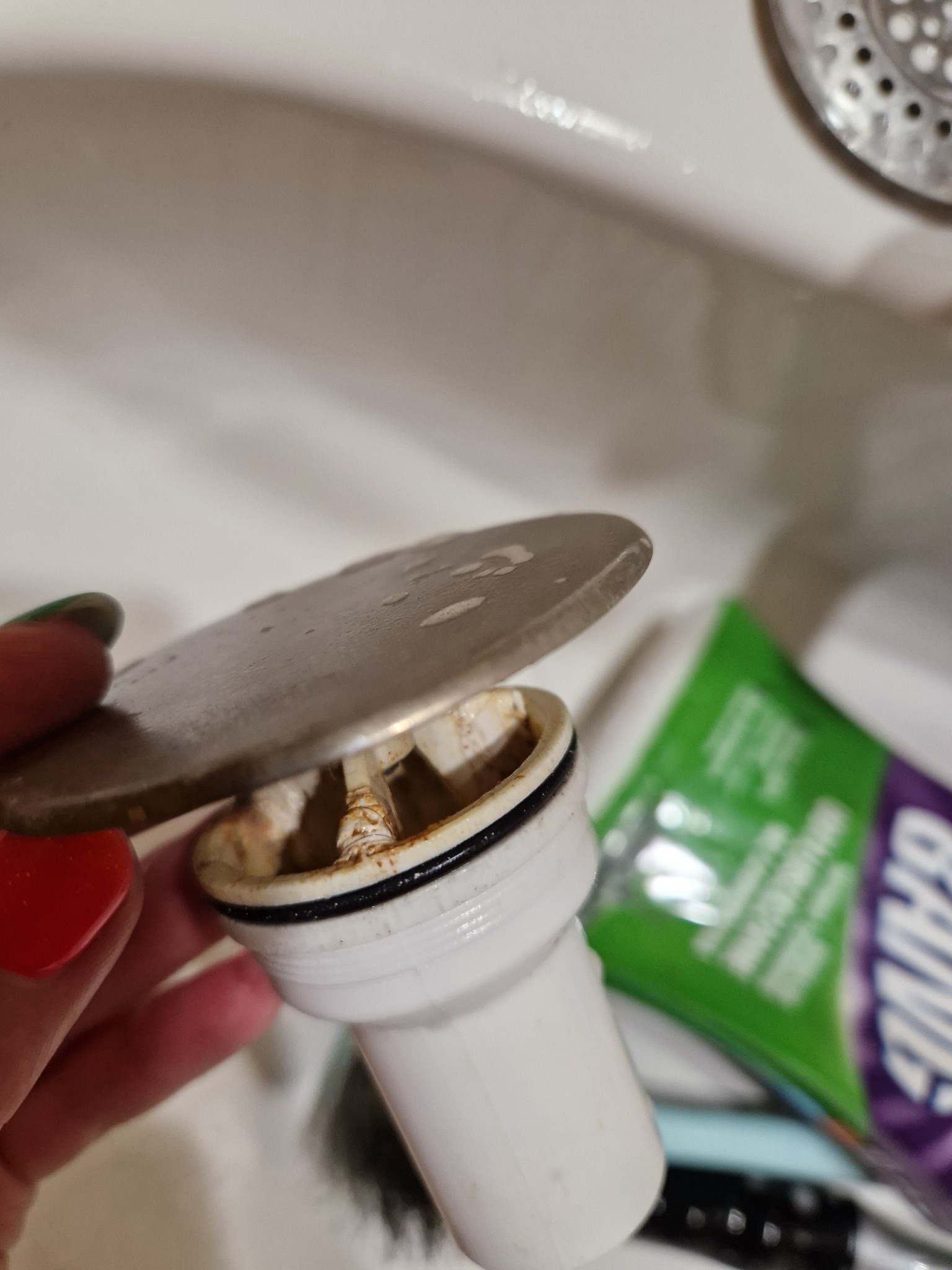 Zdemontowany, brudny syfon umywalkowy z widocznymi osadami, trzymany w dłoni, obok opakowanie środka czyszczącego