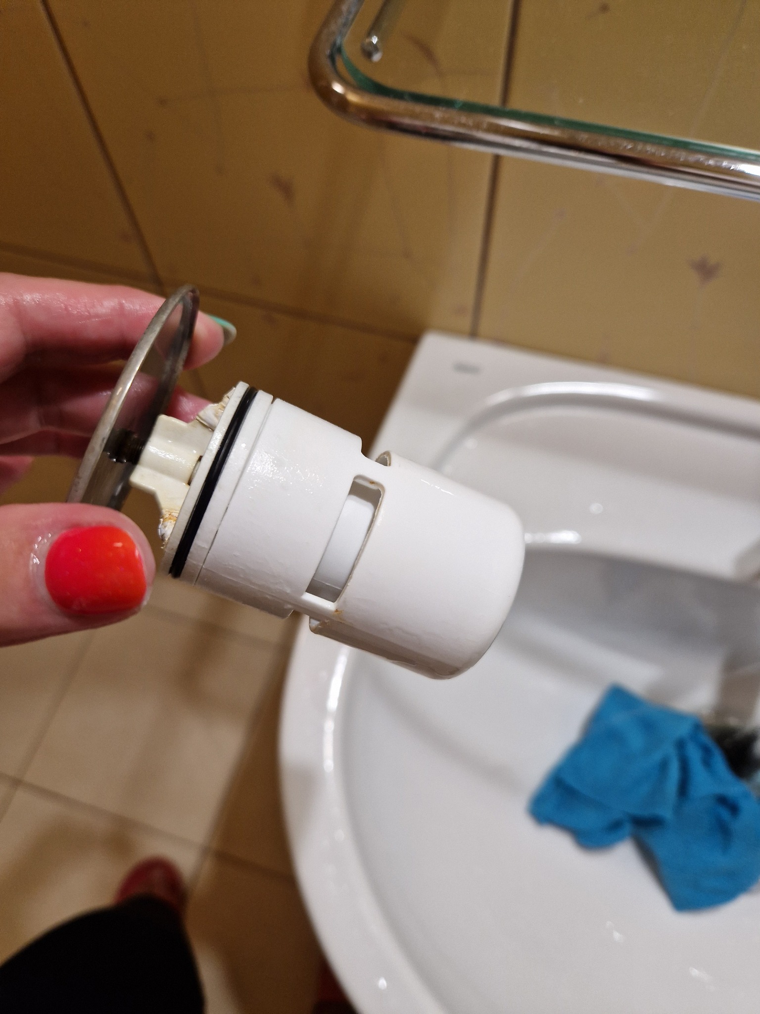 Brudny, wyjęty korek umywalkowy z osadami, trzymany w dłoni nad umywalką z niebieską ściereczką.