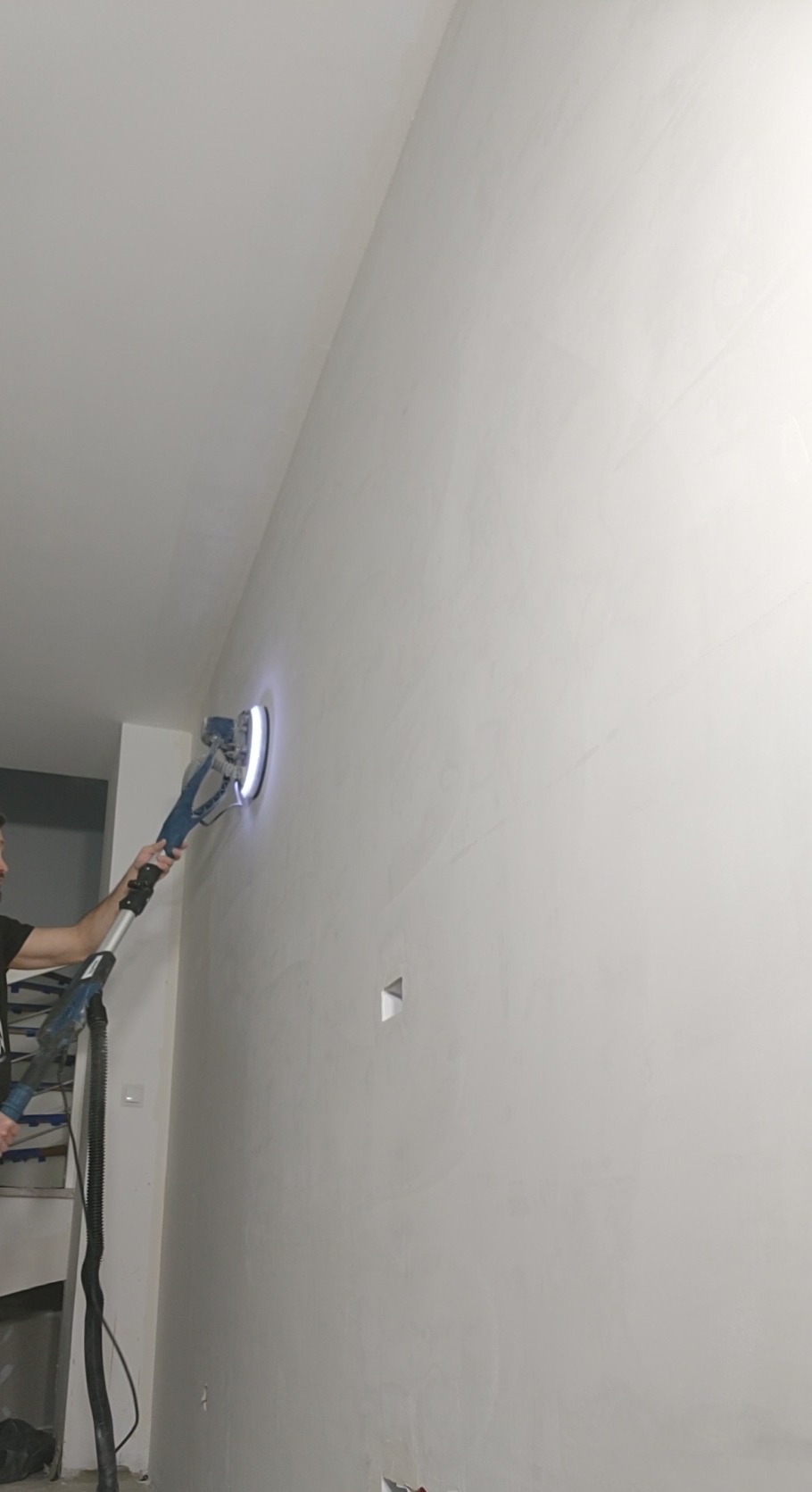 Praca szlifierką do gipsu na ścianie w trakcie wygładzania. Widoczny fragment osoby obsługującej urządzenie i przewód odkurzacza. Oświetlenie robocze szlifierki.