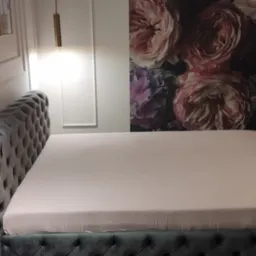 Elegancka sypialnia z szarym, pikowanym łóżkiem, nad którym wiszą złote lampy, a na ścianie widoczna jest tapeta z motywem kwiatowym.