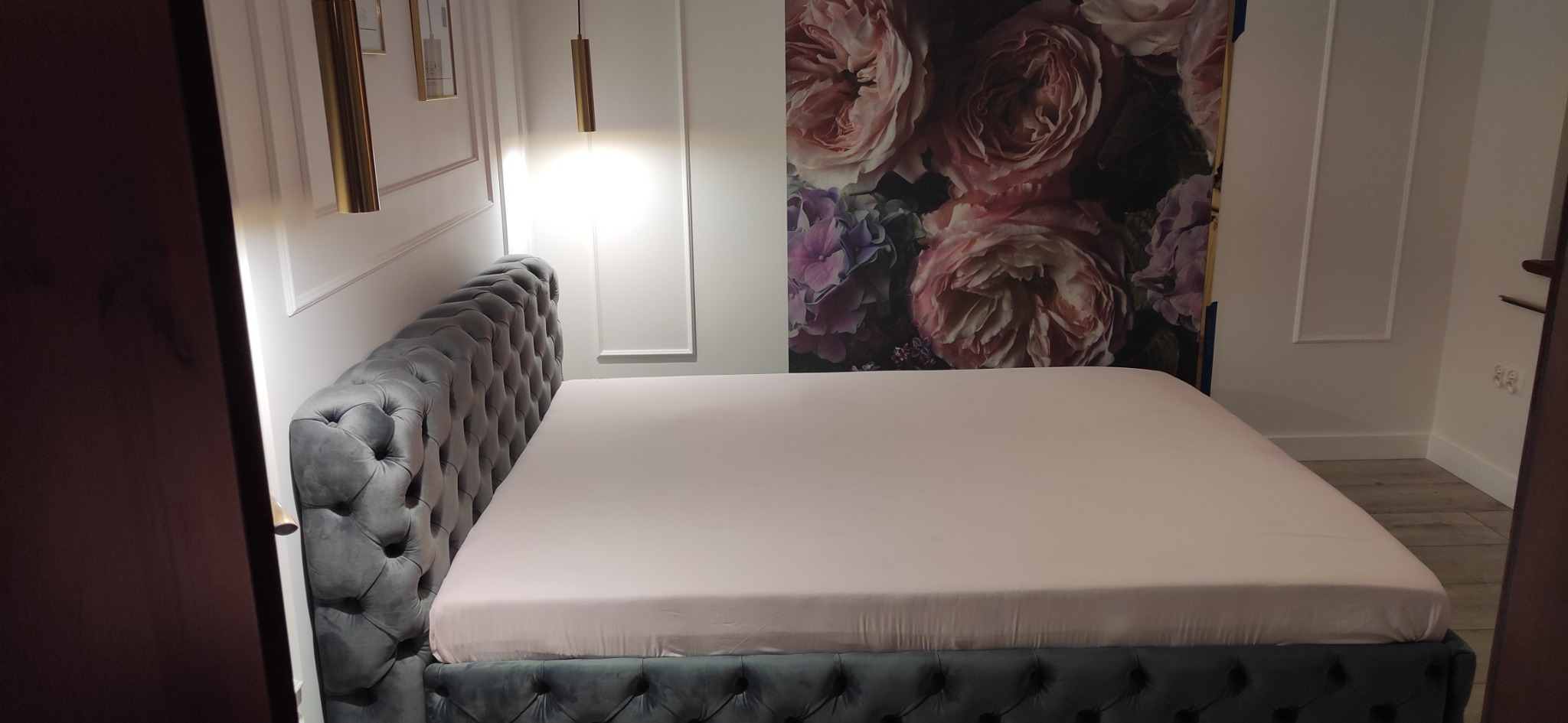 Elegancka sypialnia z szarym, pikowanym łóżkiem, nad którym wiszą złote lampy, a na ścianie widoczna jest tapeta z motywem kwiatowym.