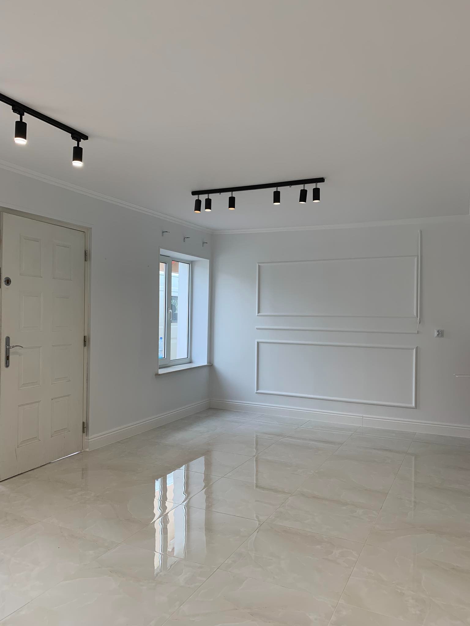 Puste, białe pomieszczenie w Tarnobrzegu po remoncie, z błyszczącą podłogą z płytek, ozdobnymi listwami na ścianie i nowoczesnym oświetleniem szynowym.