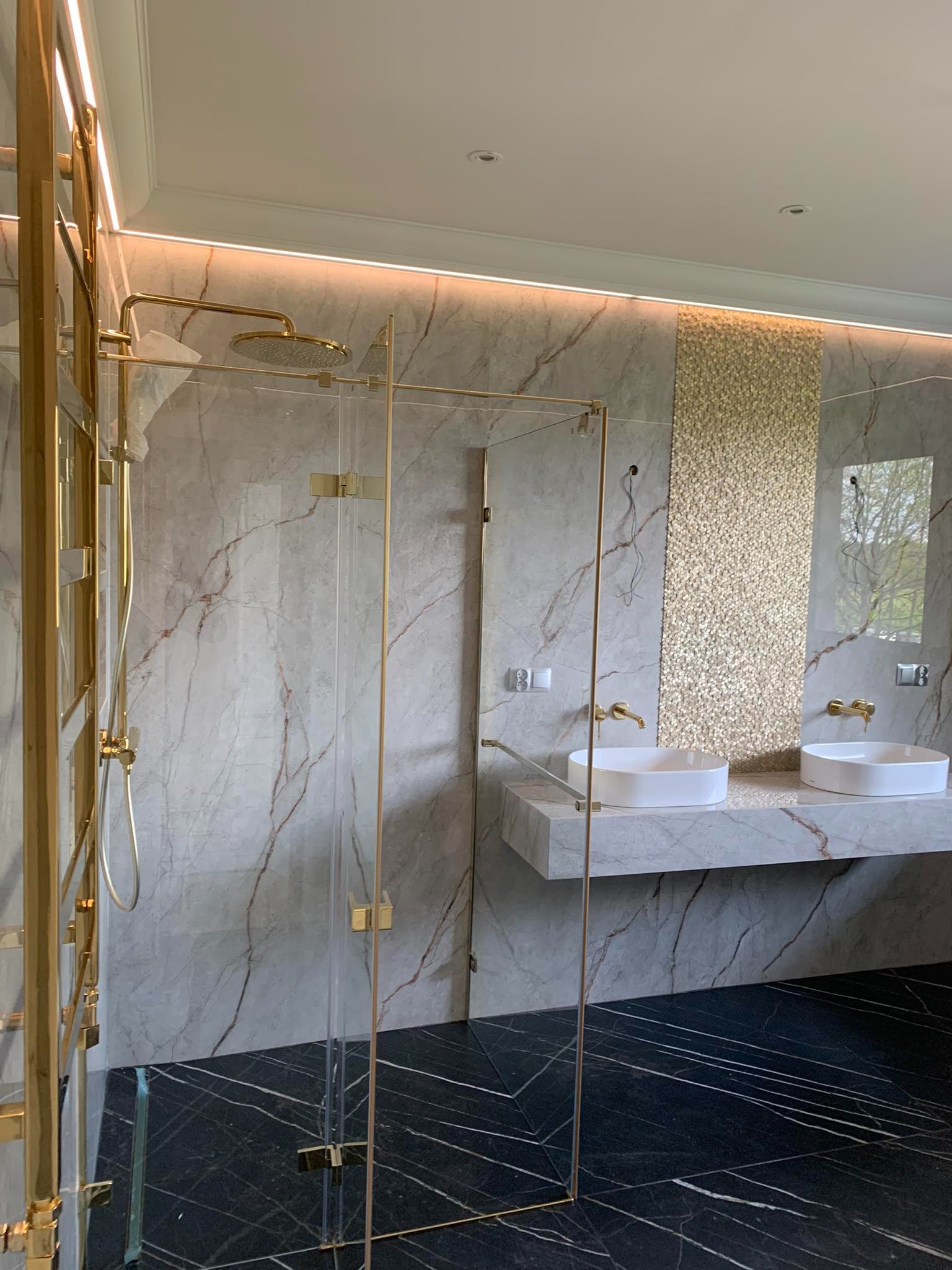 Nowoczesna łazienka z kabiną prysznicową ze złotymi elementami, umywalkami nablatowymi na marmurowym blacie i czarną podłogą z białymi żyłami.