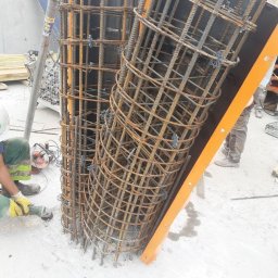 NON STOP PAO-BUD ANDRII PYLYPETS - Dwa cylindryczne szkielety z prętów stalowych, jeden w pionowej formie, drugi pochylony, widoczny pracownik w kasku i rękawicach obrabiający element szlifierką kątową, przygotowanie do zalania...