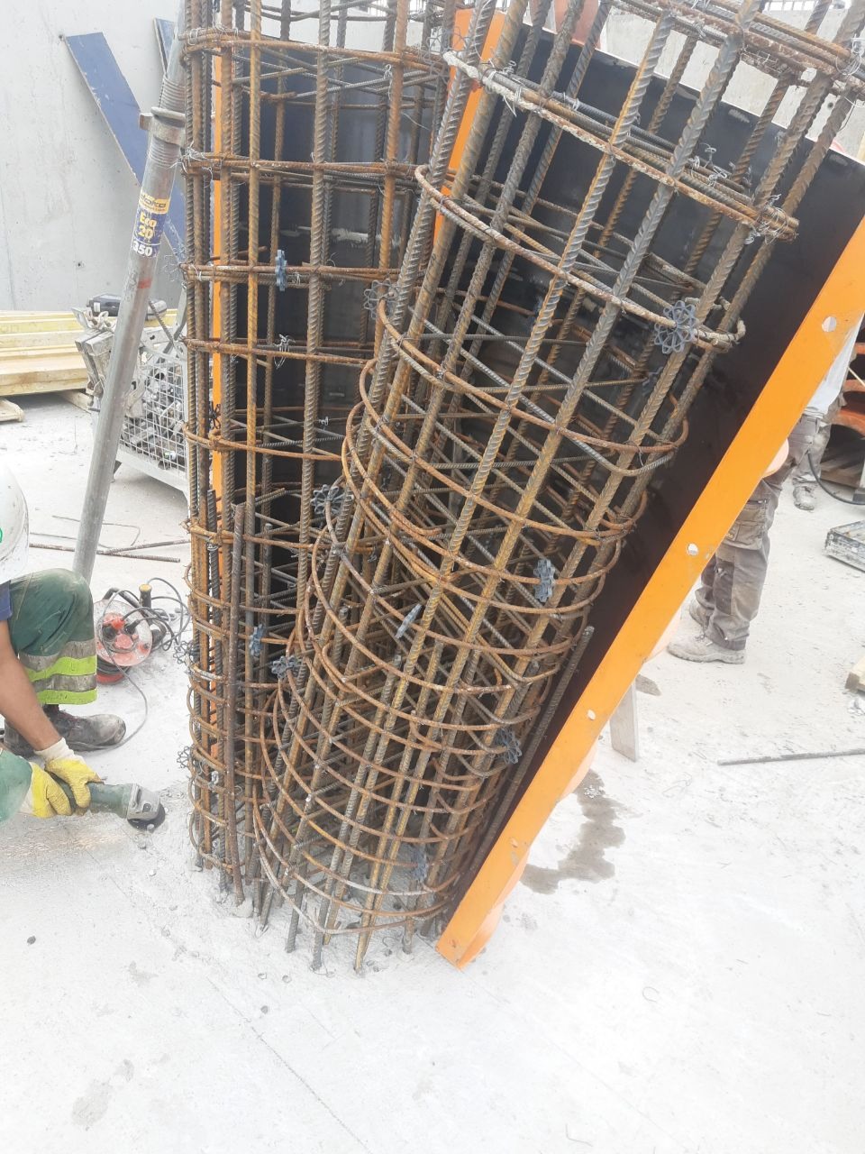 Dwa cylindryczne szkielety z prętów stalowych, jeden w pionowej formie, drugi pochylony, widoczny pracownik w kasku i rękawicach obrabiający element szlifierką kątową, przygotowanie do zalania...