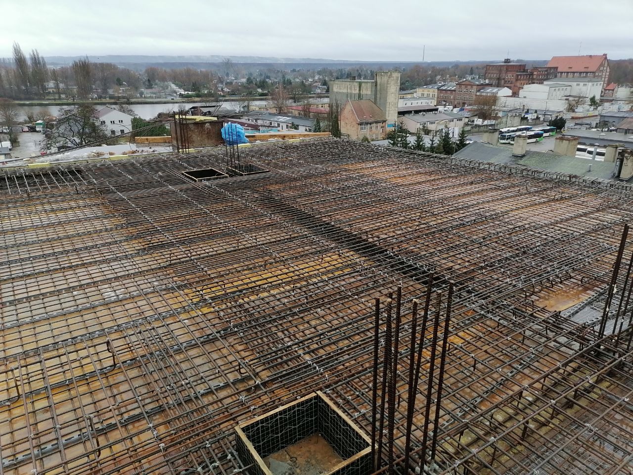 Siatka zbrojeniowa na szalunku stropu budynku, widok z góry na tle zabudowy miejskiej w pochmurny dzień.