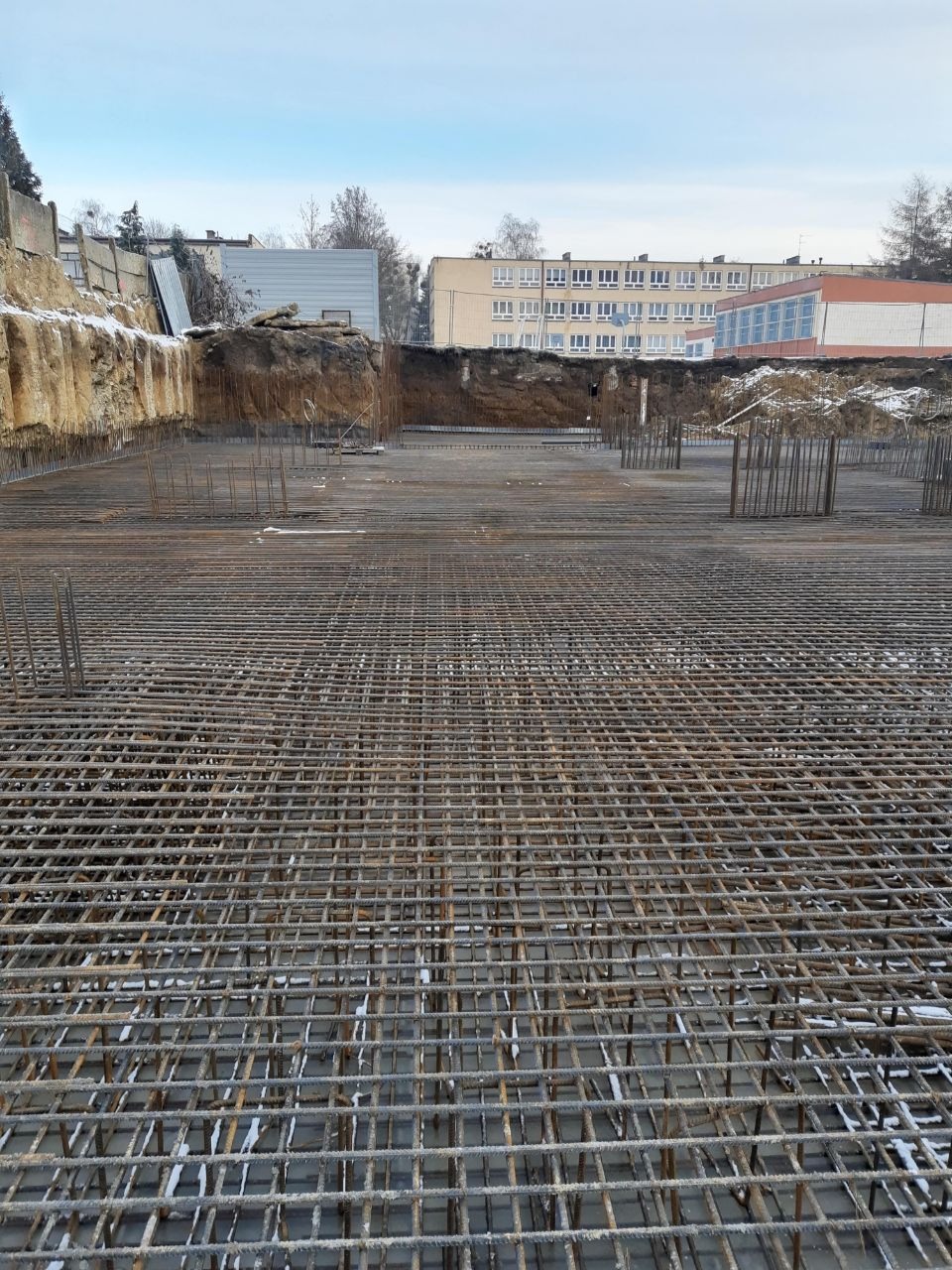 Siatka zbrojeniowa ułożona w wykopie pod fundamenty budynku, widoczne wystające pręty i ślady śniegu na betonie, w tle budynek szkolny i ziemny nasyp.