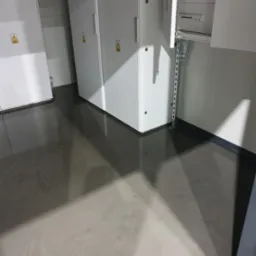 Błyszcząca, szara posadzka przemysłowa w pomieszczeniu z białymi szafami elektrycznymi i otwartą skrzynką bezpieczników.