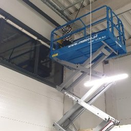 Usługi porządkowe Sylwia Szyja - Pracownik w koszu podnośnika nożycowego Mateco, na wysokości, przy oknie w hali przemysłowej z widocznymi instalacjami pod sufitem.