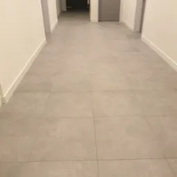 Długi korytarz wyłożony szarymi płytkami podłogowymi, z białymi listwami przypodłogowymi i drzwiami po obu stronach, oświetlony sztucznym światłem.