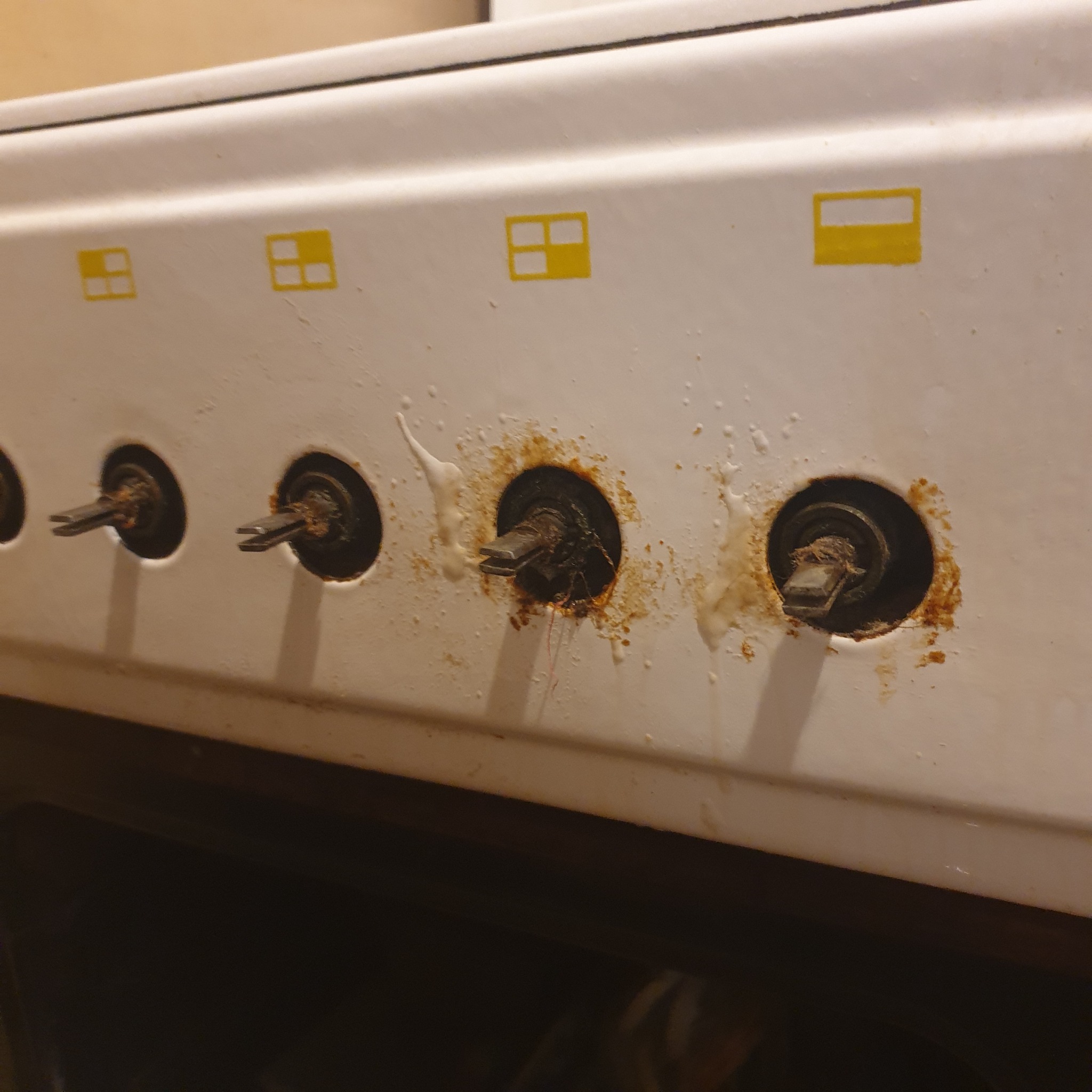 Brudny panel sterowania kuchenki gazowej z usuniętymi pokrętłami, widoczne zacieki i resztki jedzenia wokół otworów, żółte oznaczenia pól grzewczych na panelu.